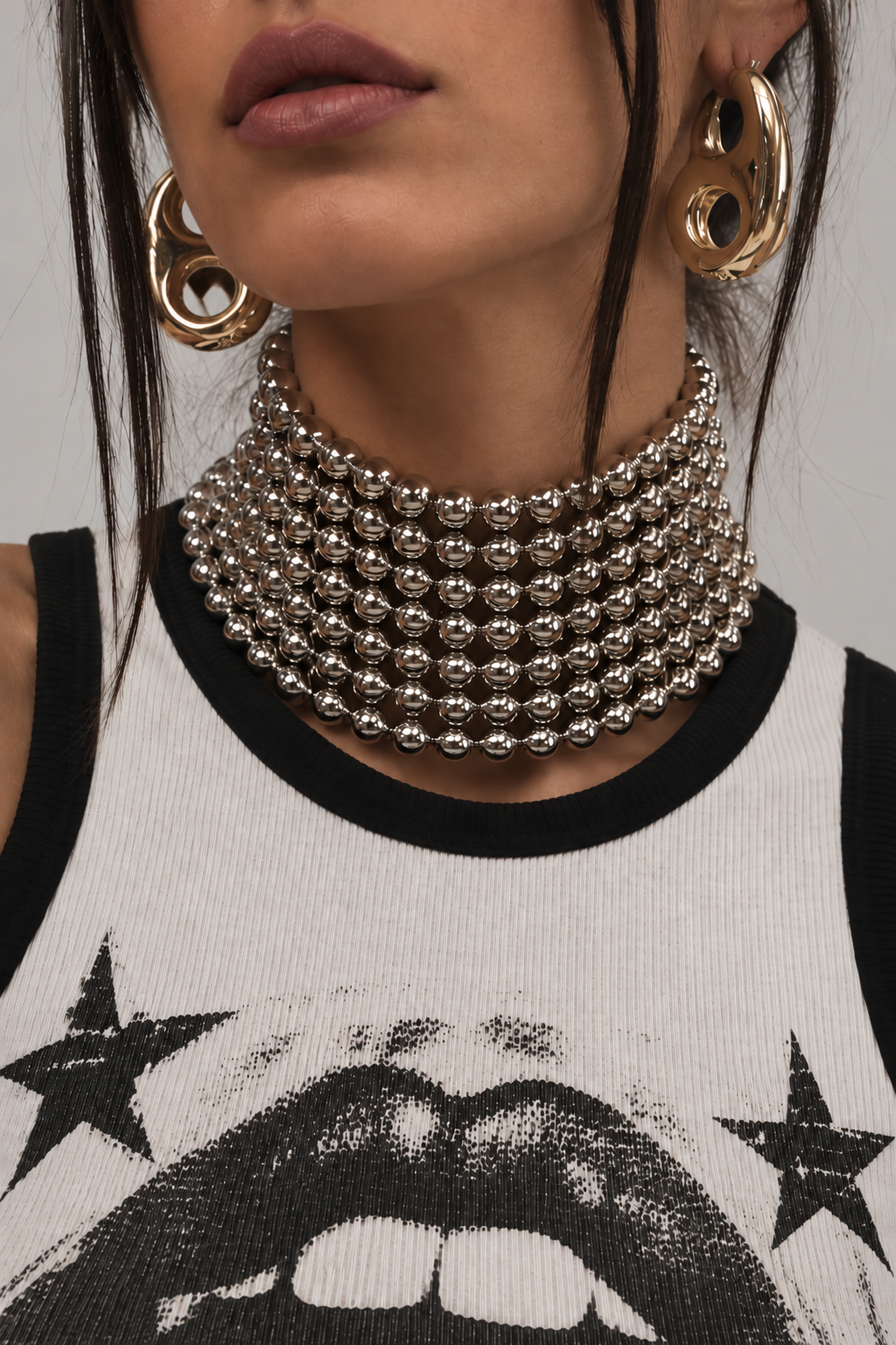 A woman wears large gold hoop earrings and the FULL FORCE CHOKER IN SILVER—a chunky, beaded statement necklace—paired with a sleeveless black and white graphic top featuring a mouth and stars, creating an ideal concert look. Her hair partially obscures her face.