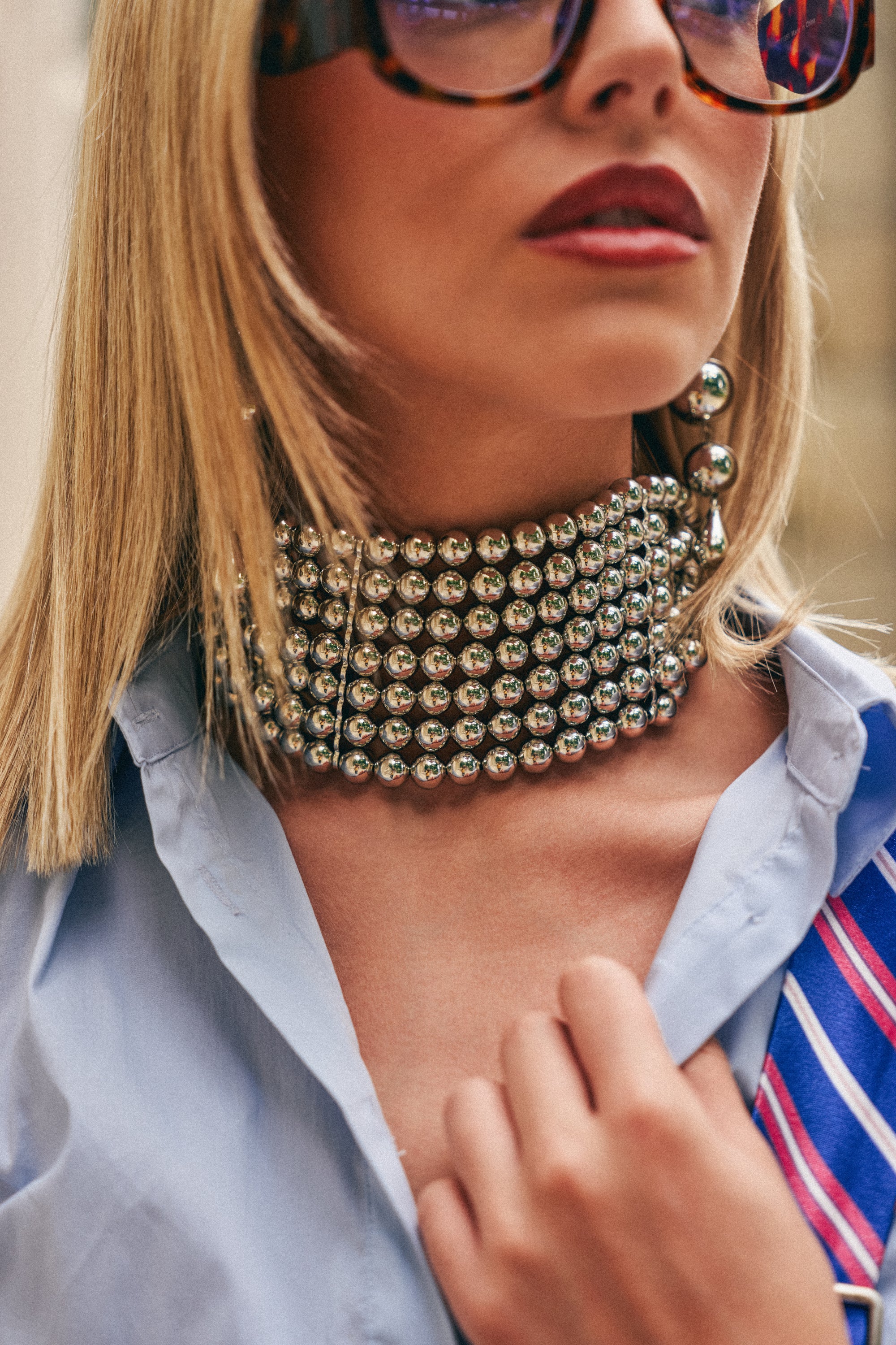 A woman wears oversized sunglasses, a light blue shirt while holding her collar, and the FULL FORCE CHOKER IN SILVER—a bold, multi-layered beaded statement necklace. Her lips are painted a deep red and she sports one dangling earring for an effortlessly chic concert-ready look.