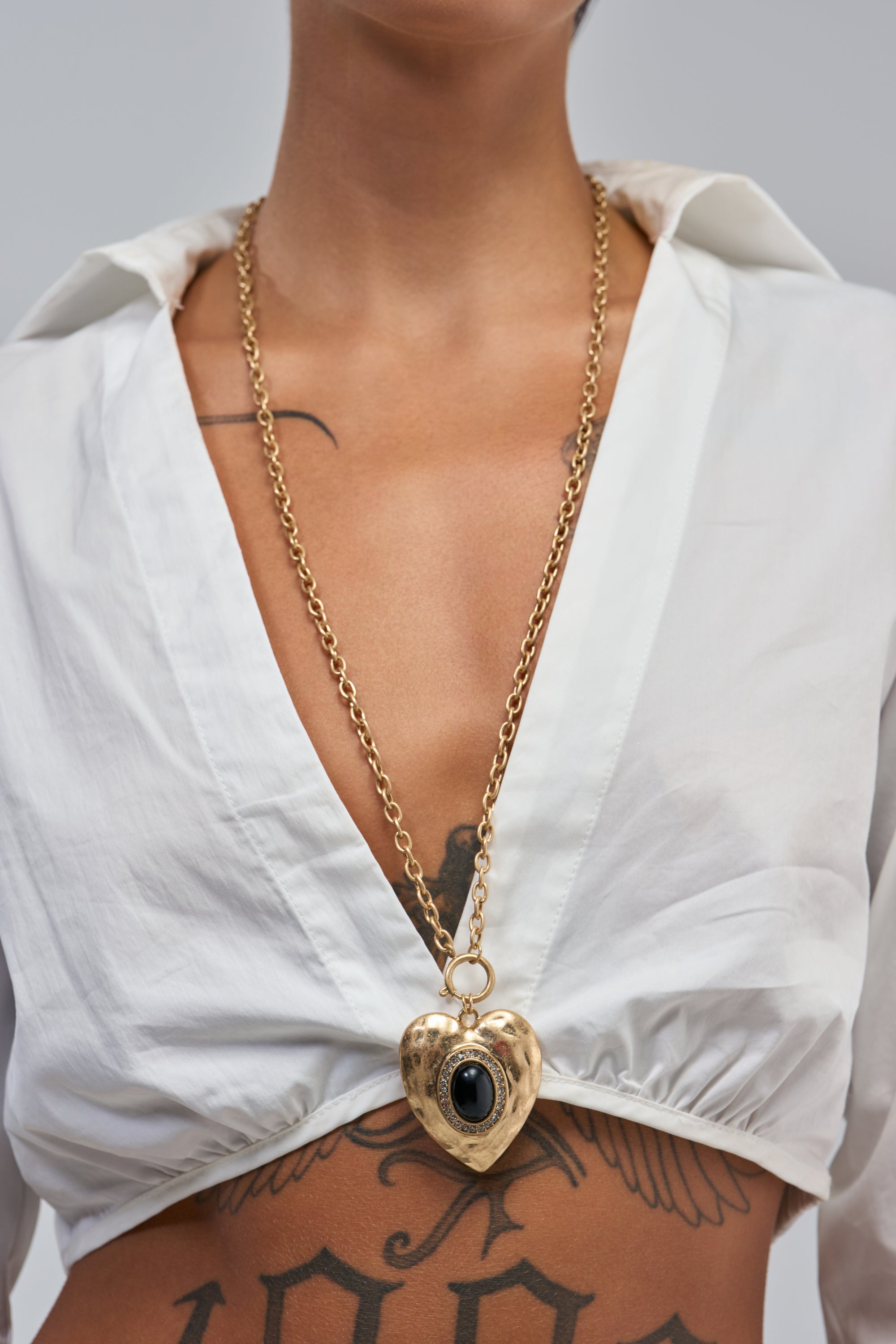 A person in a white deep V-neck top wears the ANTIQUE HEART PENDANT NECKLACE—featuring a gold heart with a black stone on a long chain—while black tattoos are visible on their chest and stomach.