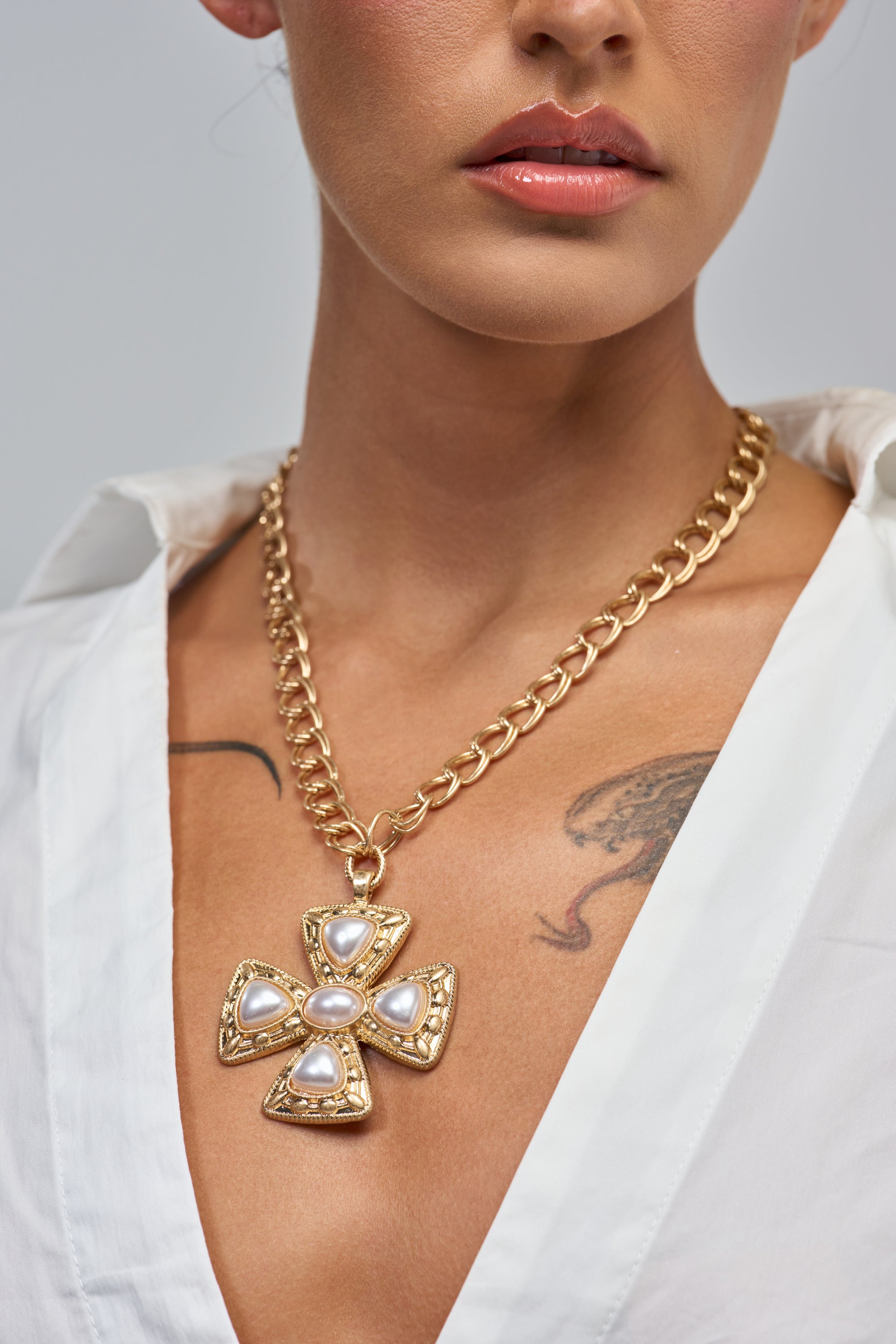 A woman in a white shirt wears the ANTIQUE CROSS PENDANT NECKLACE—a chunky double chain with a large gold cross adorned with pearls. A serpent tattoo is partly visible on her chest.