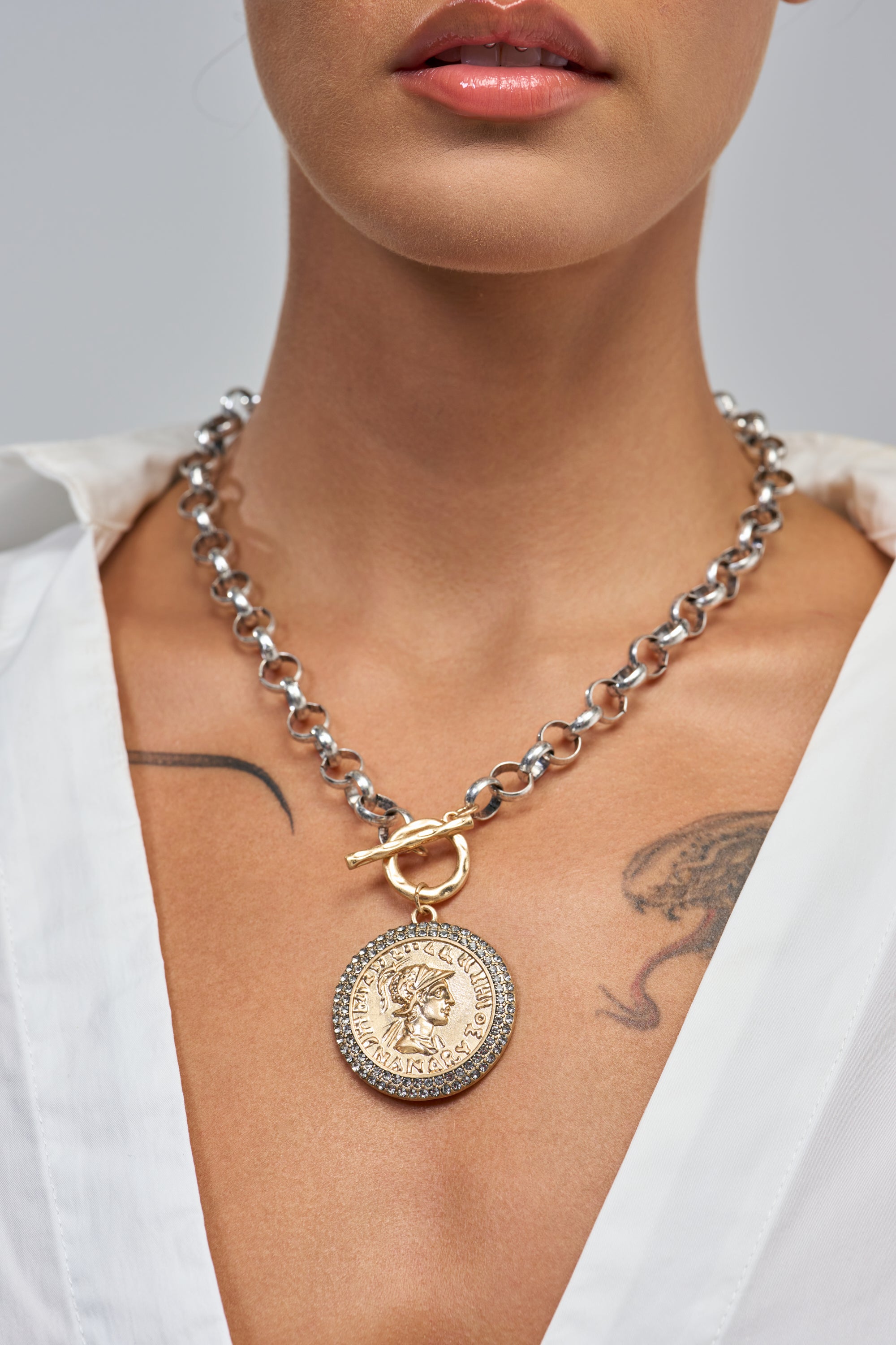 A person wears the ANTIQUE MEDALLION PENDANT NECKLACE—a chunky silver chain with a medallion—over an open-collared white shirt, revealing part of a chest tattoo.