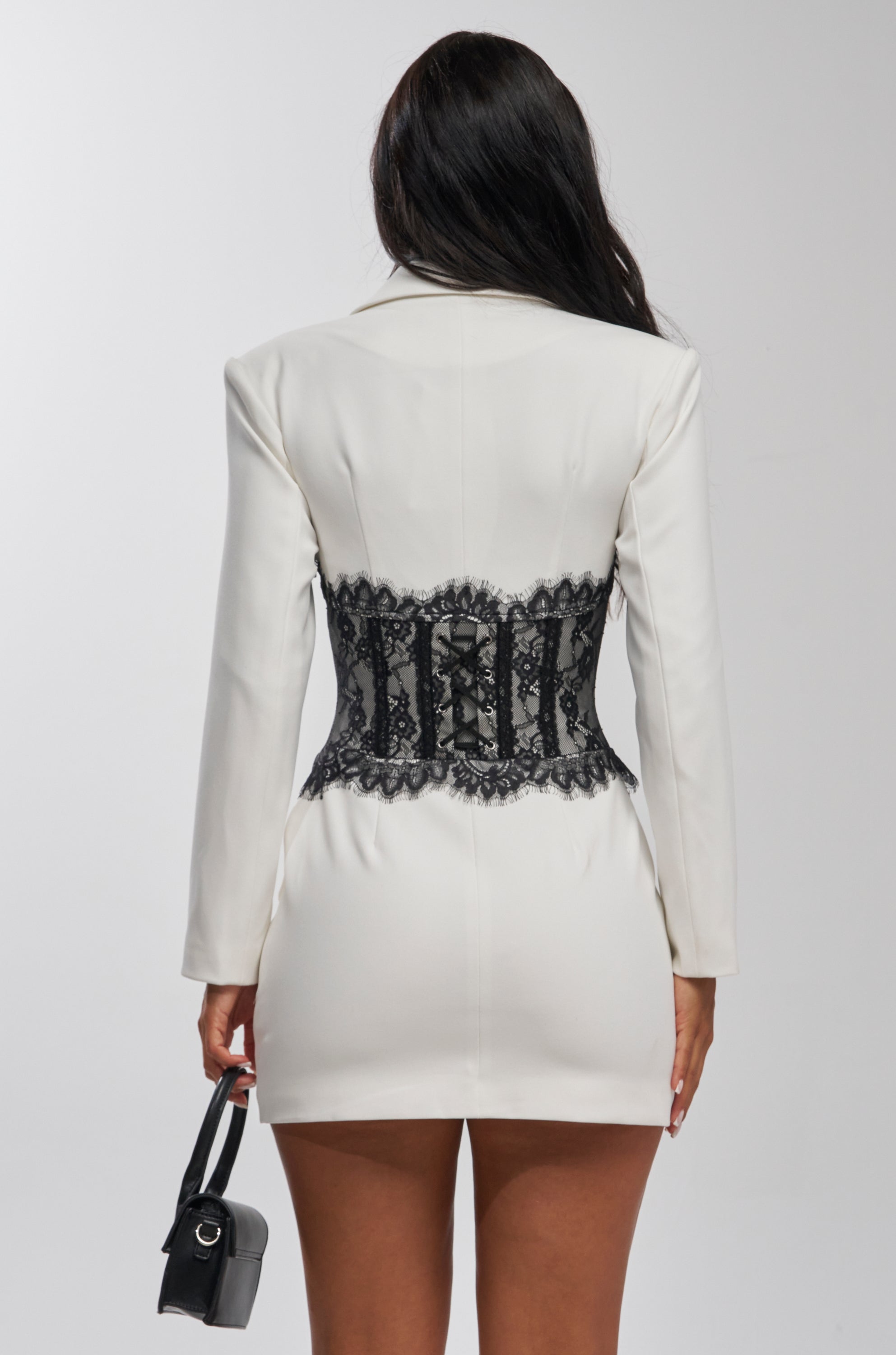A woman stands facing away, wearing the STAR BEHAVIOR LACE BLAZER DRESS IN BLACK and holding a small black handbag—a stunning cocktail party look against a light, plain background.