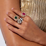 A close-up of a person's hand adorned with the stunning TAKE ME ON A TRIP RING SET, featuring statement rings with an emerald, a ruby, and a striking black-and-white design. Set against gold, mesh-like fabric, these ornate rings gleam with their gemstone embellishments as the elegantly positioned hand exudes vacay vibes.