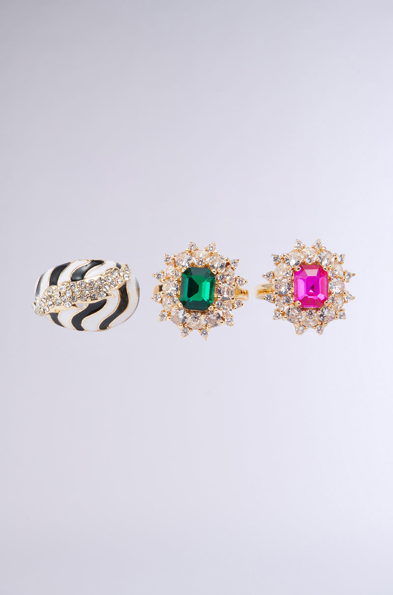 A photo features the TAKE ME ON A TRIP RING SET. From left to right: a ring adorned with a black and white zebra stripe pattern and rhinestones, a ring featuring a large green gemstone with small diamonds around it, and a ring showcasing a large pink gemstone also encircled by small diamonds. 1