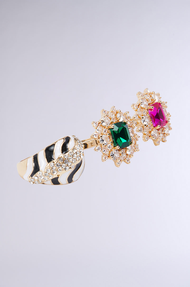 The TAKE ME ON A TRIP RING SET includes a gold ring with a distinctive black and white patterned band and two prominent gemstones. One gem is an emerald-cut green stone encircled by smaller clear stones, while the other features an emerald-cut pink stone with a similar halo of clear stones — ideal for elevating your vacation style with a touch of gemstone elegance. 2