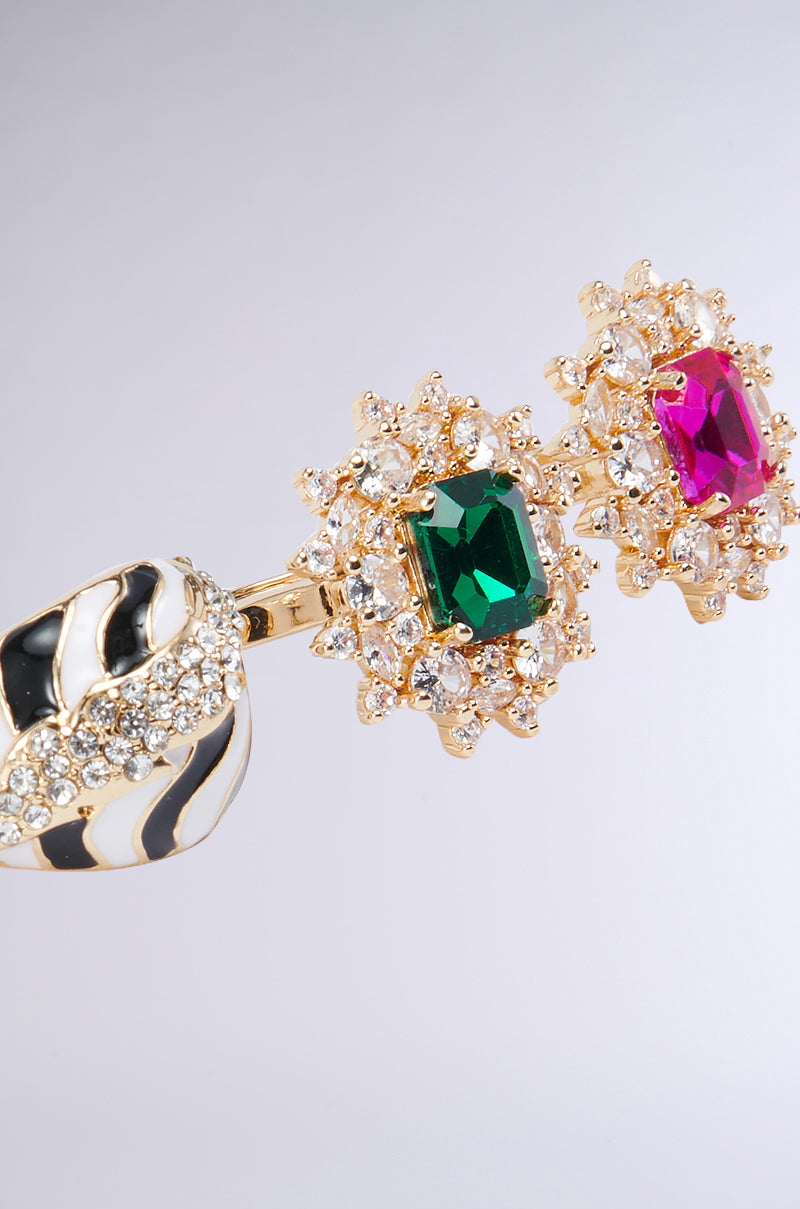 A close-up image of the TAKE ME ON A TRIP RING SET showcases rhinestone-encrusted embellishments. It features a green rectangular gemstone surrounded by clear crystals on one side and a pink rectangular gemstone, also encircled by crystals, on the other. The band boasts a black-and-white striped design with additional clear crystals. 3