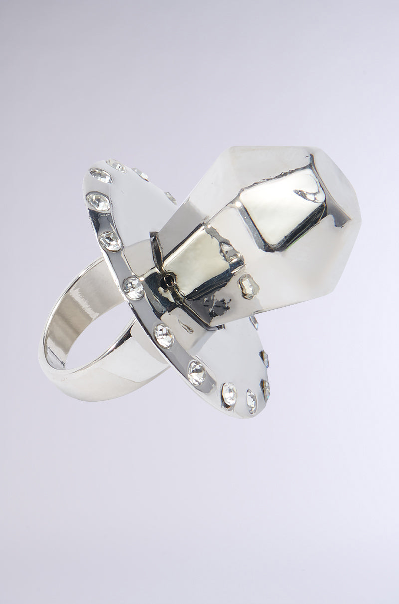 Introducing the SUCK ON THAT COCKTAIL RING: a bold UFO-shaped design showcasing a prominent central faceted gemstone. This piece is crafted with a round base embellished with rhinestone accents mimicking lights and complemented by a structured band, set against a plain gradient background transitioning from light gray to white.