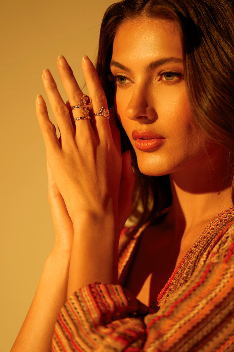 A woman with long brown hair and subtle makeup clasps her hands near her face, showcasing the CROSS YOUR HEART RING SET. Her textured, colorful garment complements the faux gold rings as warm golden lighting casts soft shadows on her face. 0