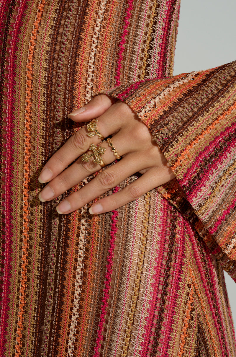 A close-up shows a hand wearing the CROSS YOUR HEART RING SET with intricate designs. The hand rests on a colorful fabric featuring vertical stripes in brown, orange, pink, and cream, with a slightly flared cuff. 2