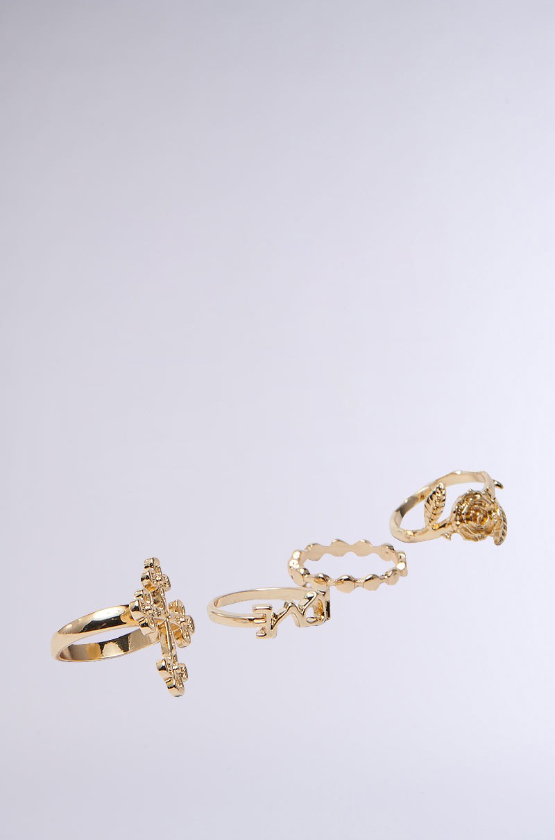 The CROSS YOUR HEART RING SET features three faux gold rings: a delicate rose, bold geometric shapes, and an ornate key, all uniquely designed and elegantly displayed against a simple backdrop. 3