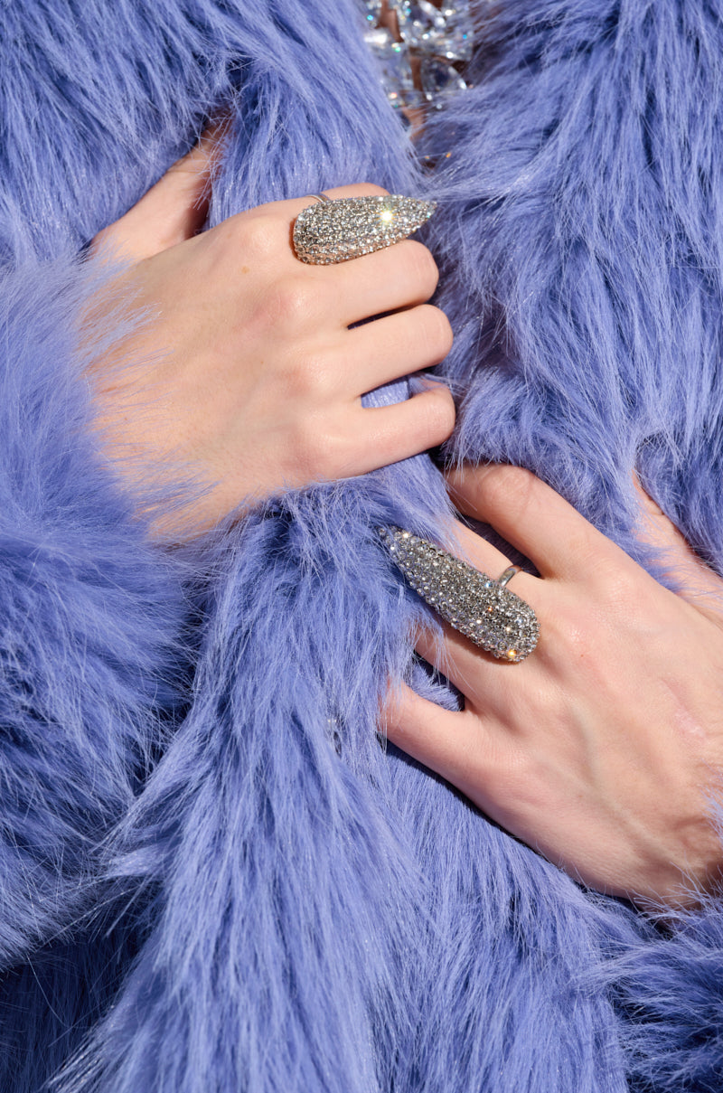 A person wearing a soft, light purple faux fur coat, with hands adorned in glittering adjustable rings from the CHECK OUT MY CLAWS RING SET. The coat and rings embellished with silver rhinestones create a luxurious and stylish combination.