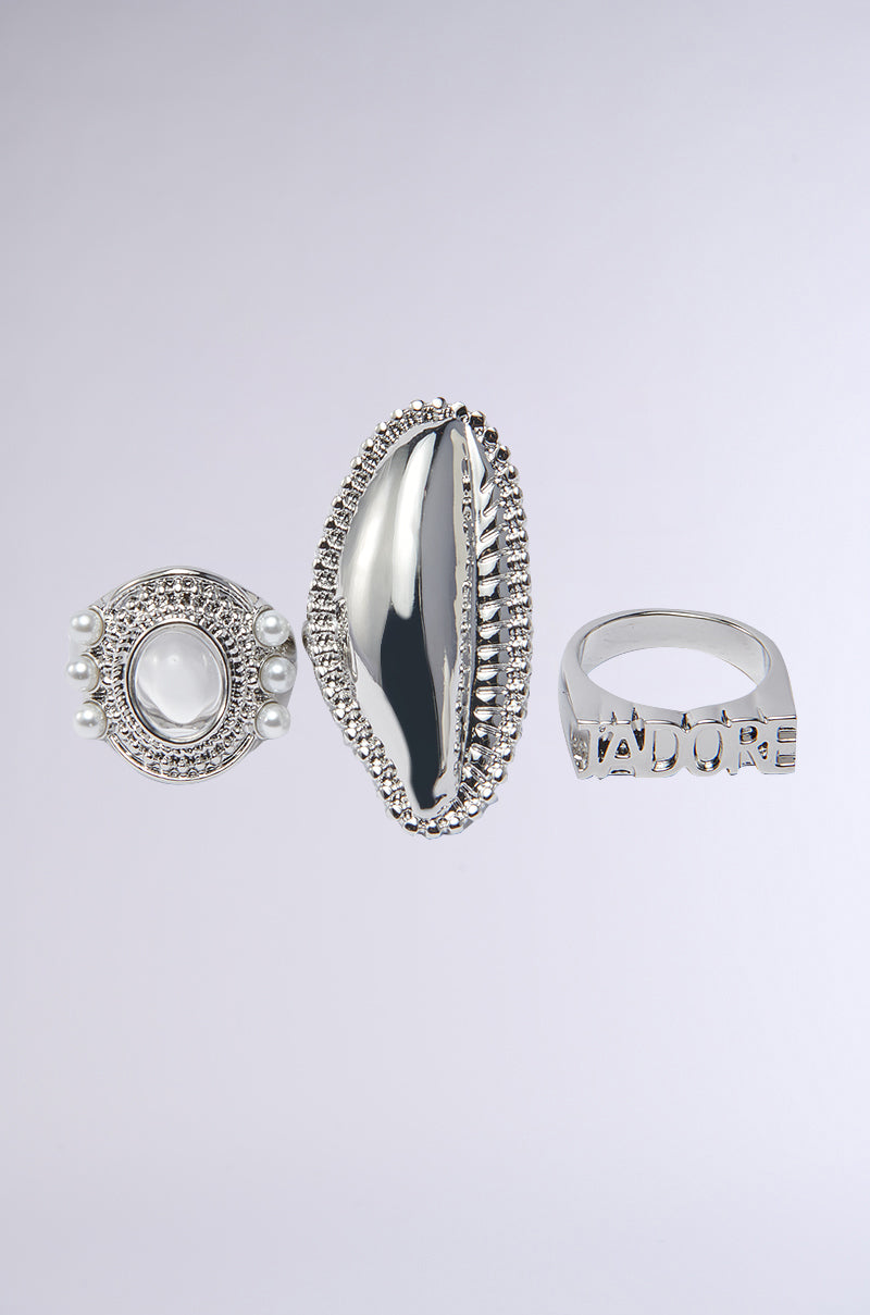 The RIRI RING SET features three silver-tone rings: one with a central oval stone and pearl accents, one elongated with texture, and one engraved with J'ADORE lettering, displayed on a light gray background.