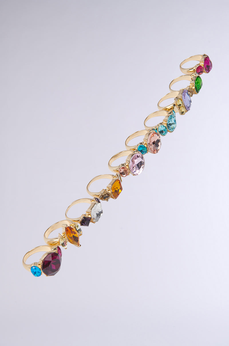 A diagonal row of gold-tone rings set with oversized blue, pink, purple, green, red, and yellow gemstones showcases the SUMMER VIBES RING SET on a light gray background. 2