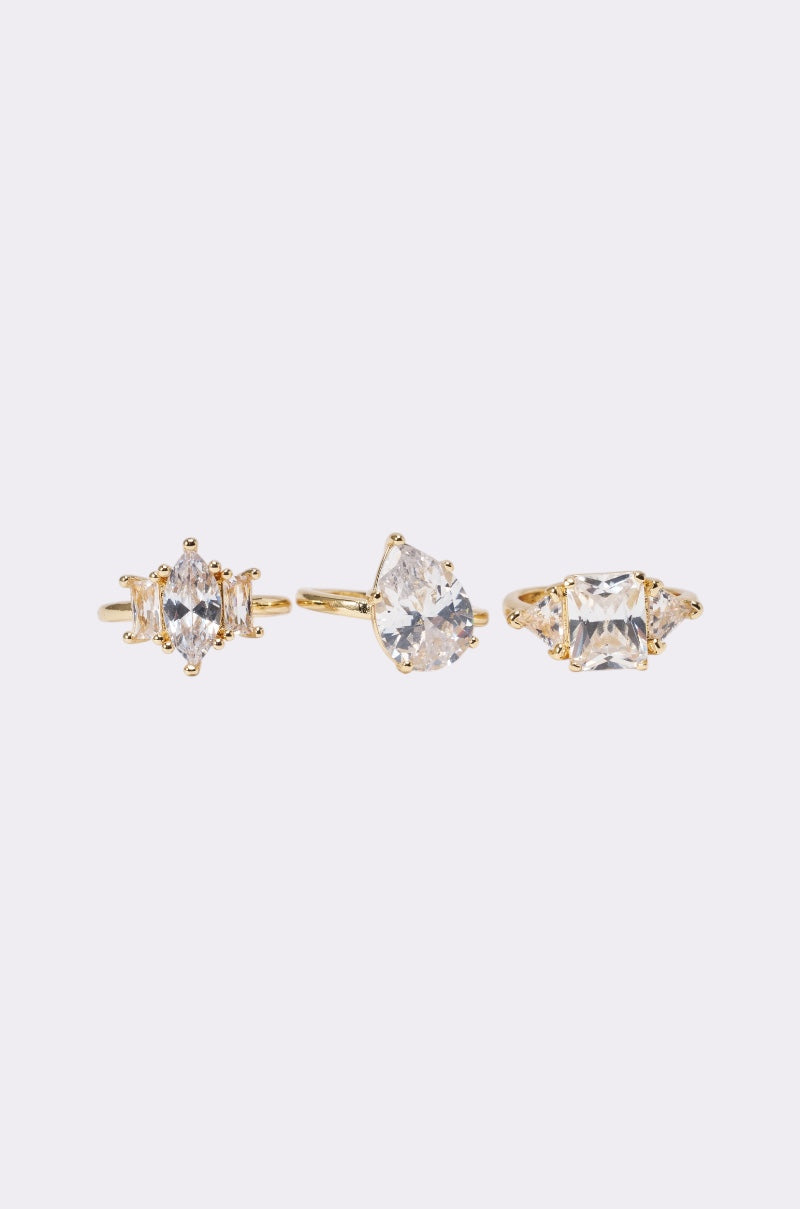 The TOP TIER RING SET features three gold-tone rings with large clear gemstones in oval, heart, and square shapes, adorned with crystal accents and displayed side by side on a plain light background to showcase their elegance. 1