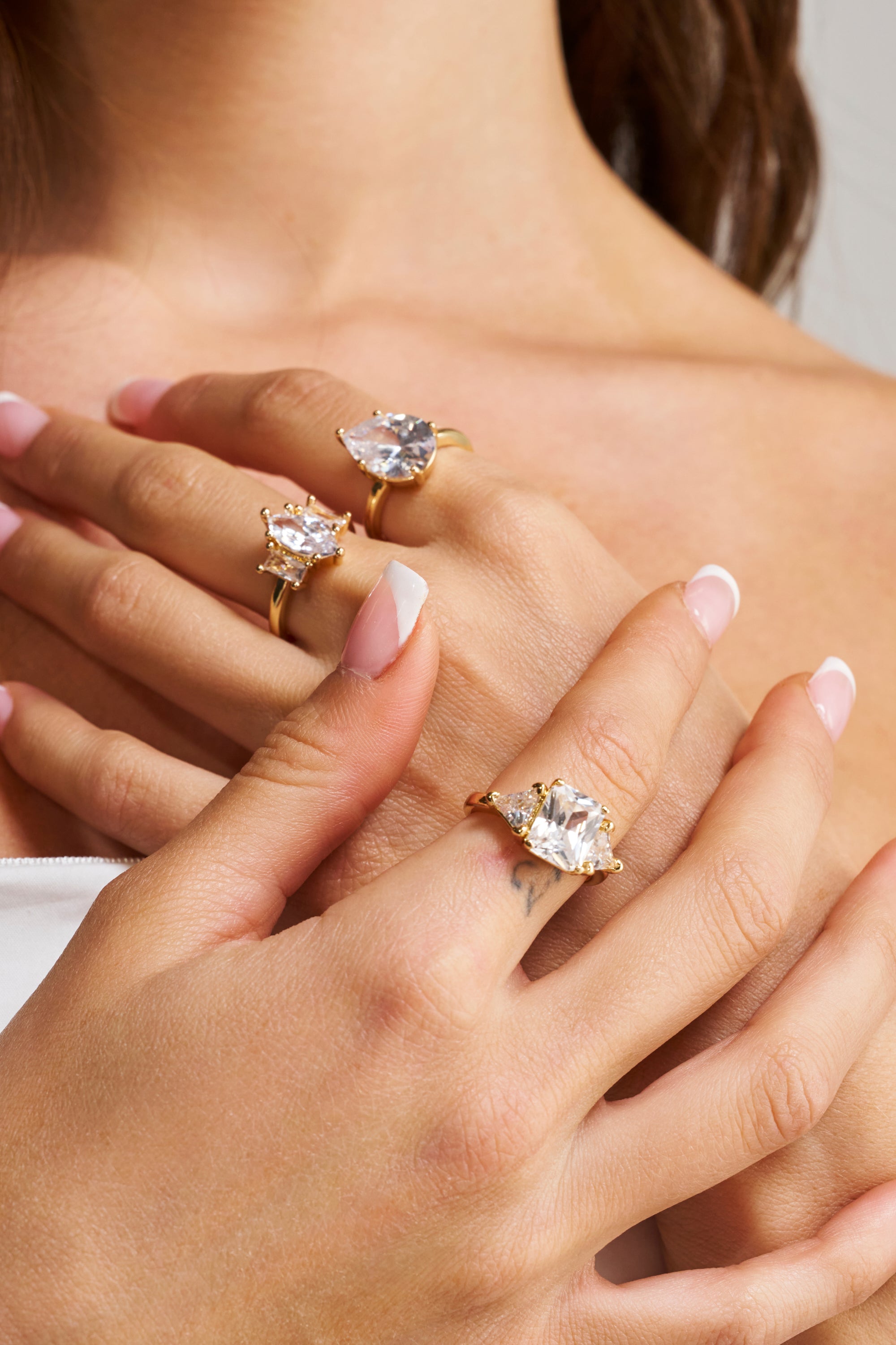 A close-up shows a woman's manicured hands wearing the TOP TIER RING SET, featuring gold-tone bands with large, clear gemstones. The elegant formal jewelry contrasts beautifully with her light pink nails and smooth skin.