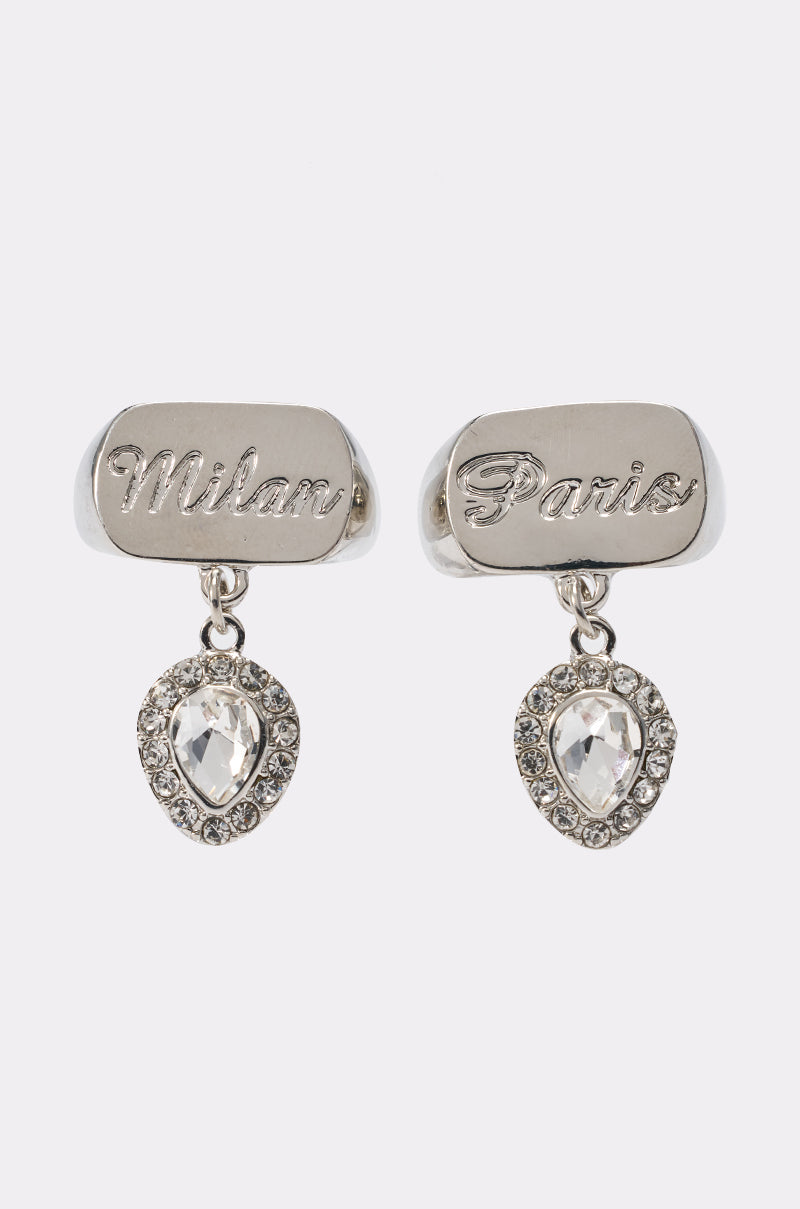The IN YA CITY RING SET features silver rings engraved with "Milan" and "Paris," each adorned with teardrop crystal charms surrounded by small stones, creating an elegant, sparkling look beneath the inscriptions. 1