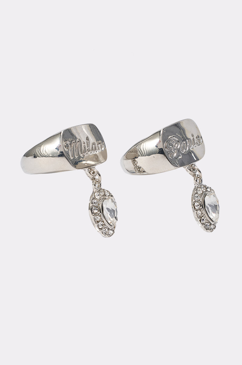 The IN YA CITY RING SET features two silver rings with engraved nameplates, "Milan" and "Paris," each adorned with dangling oval crystal charms bordered by small crystals. 2