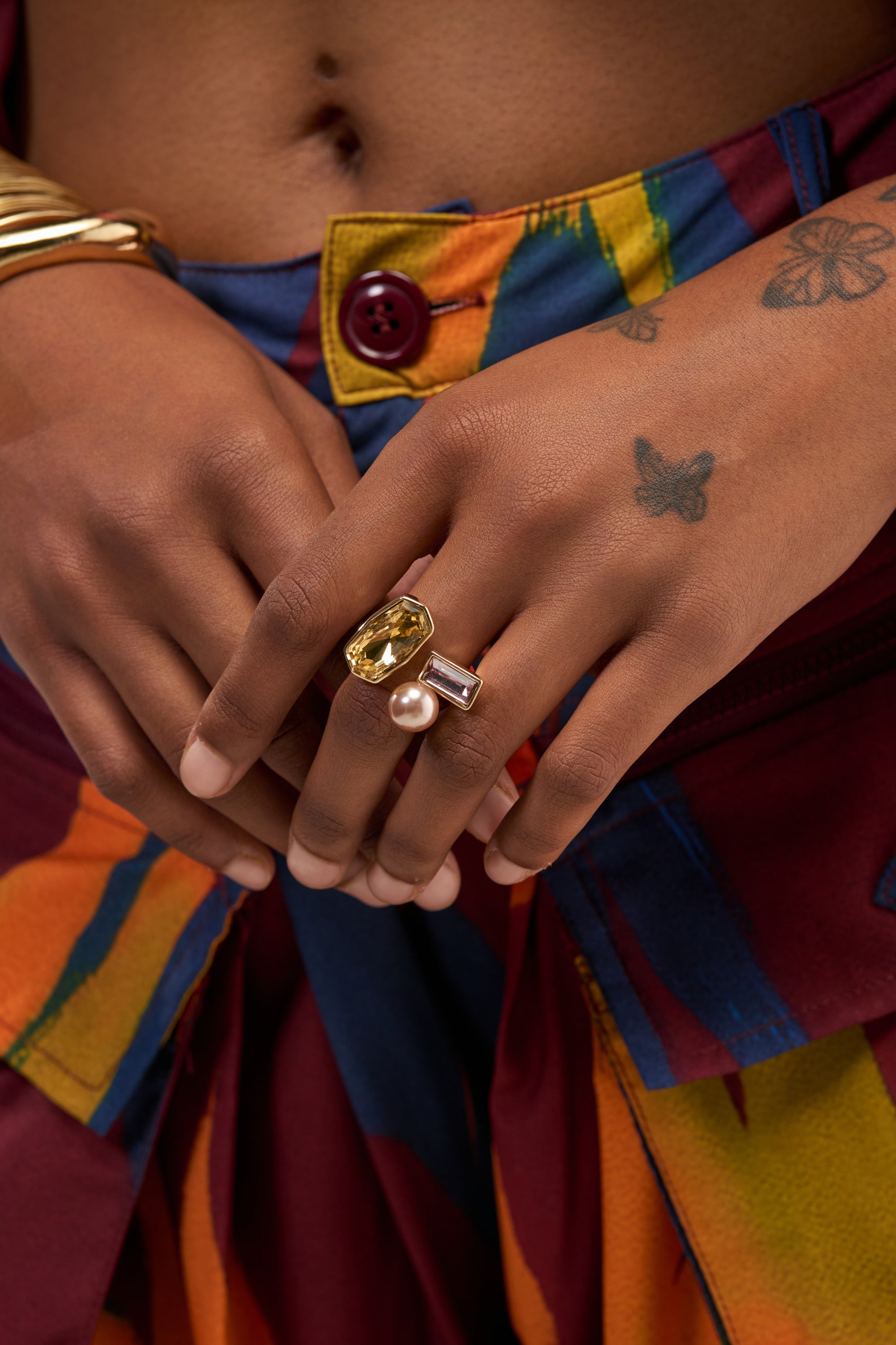 A person with butterfly tattoos on their hand wears colorful pants and showcases an eye-catching array of rings—among them, a pearl ring, a yellow gem, a baguette stone, and the distinctive WHISKEY CRYSTAL RING. Their hands are clasped near their waist, highlighting the unique statement piece adorning their fingers.