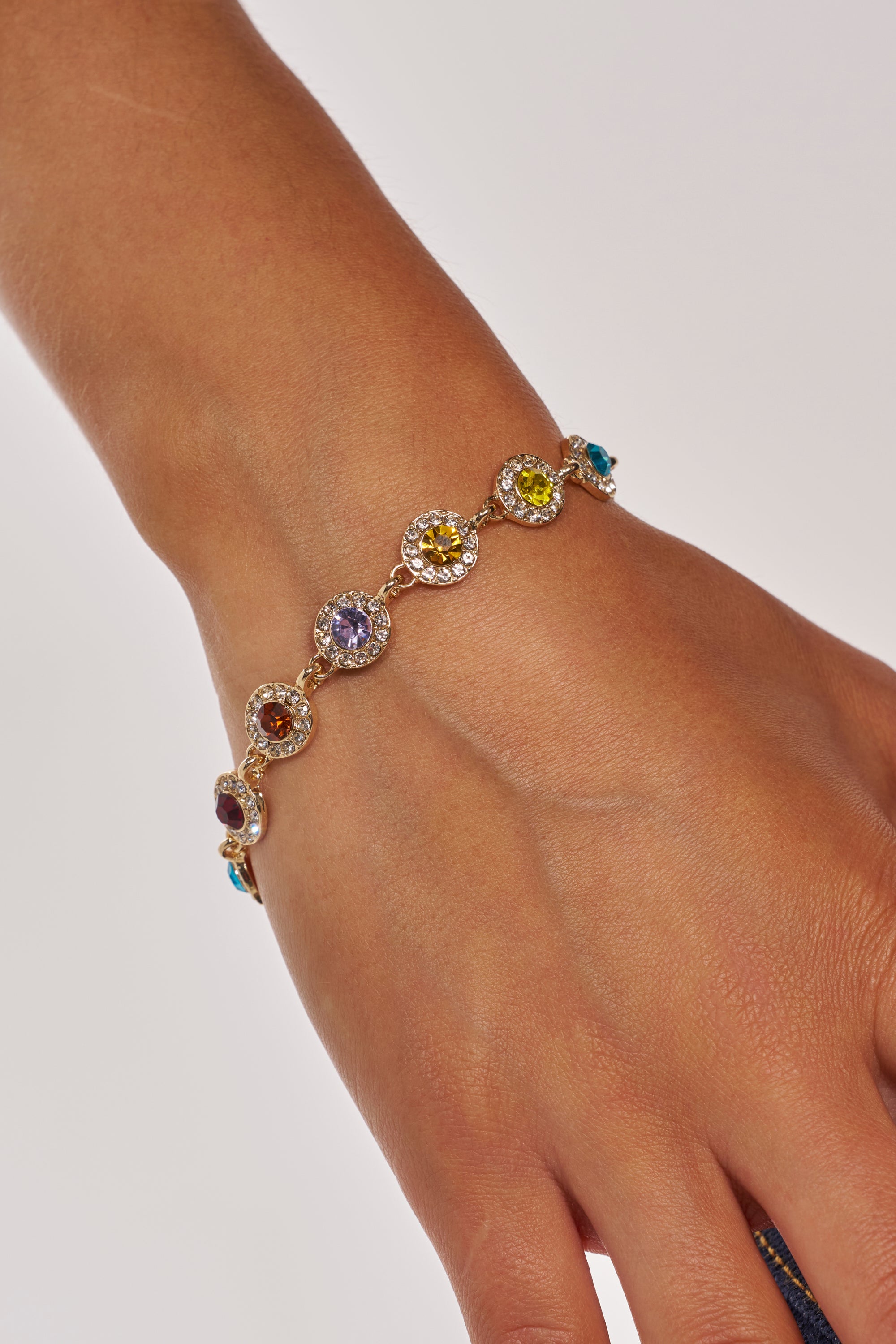 A close-up of a COME CORRECT NECKLACE, featuring gold-tone metal set with round, multicolor faceted stones—purple, yellow, and blue—sparkling vividly against a light background. 3