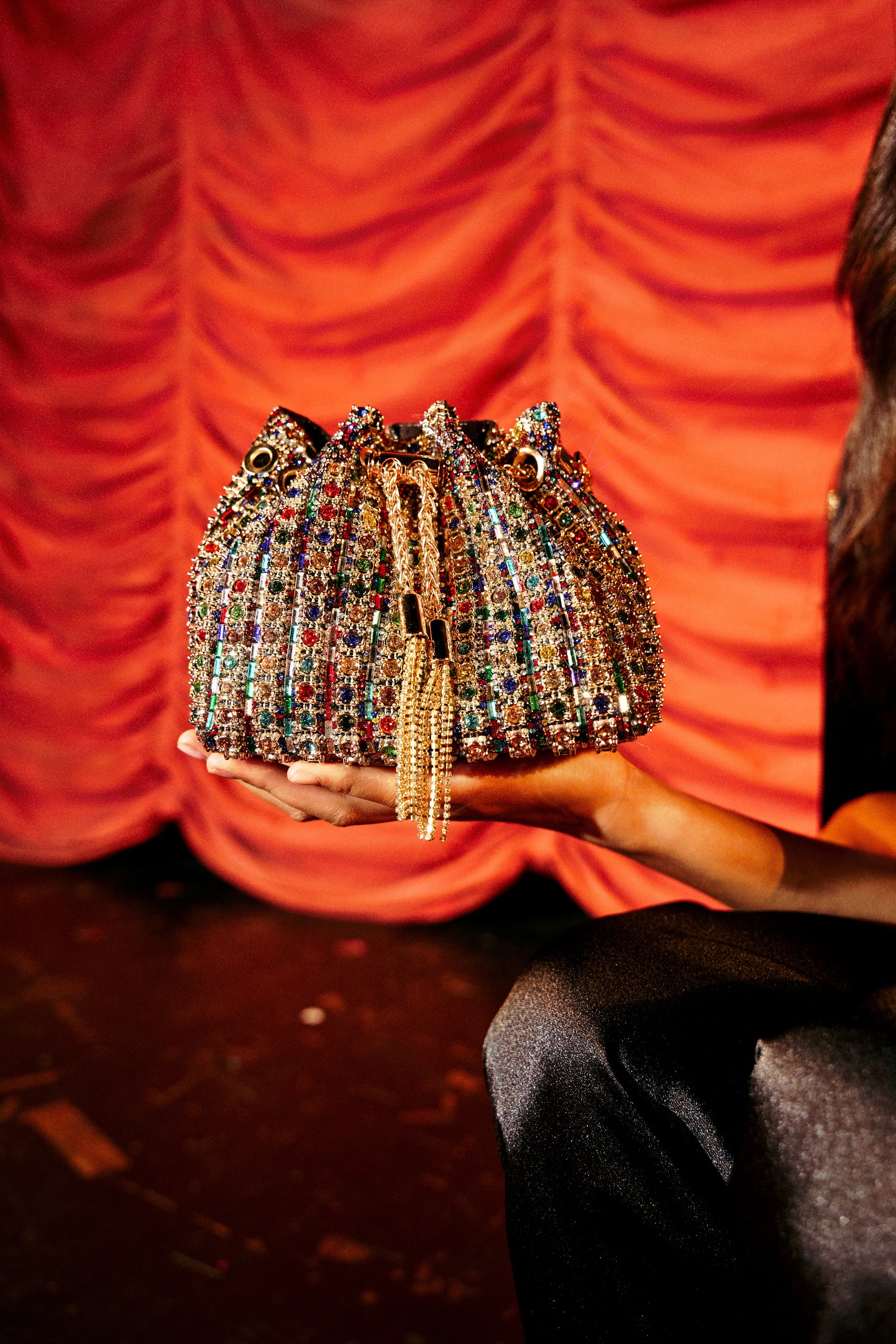 A hand holds the GEGE BLING PURSE, a colorful rhinestone bag with gold chain tassels, set against dramatic red curtains.