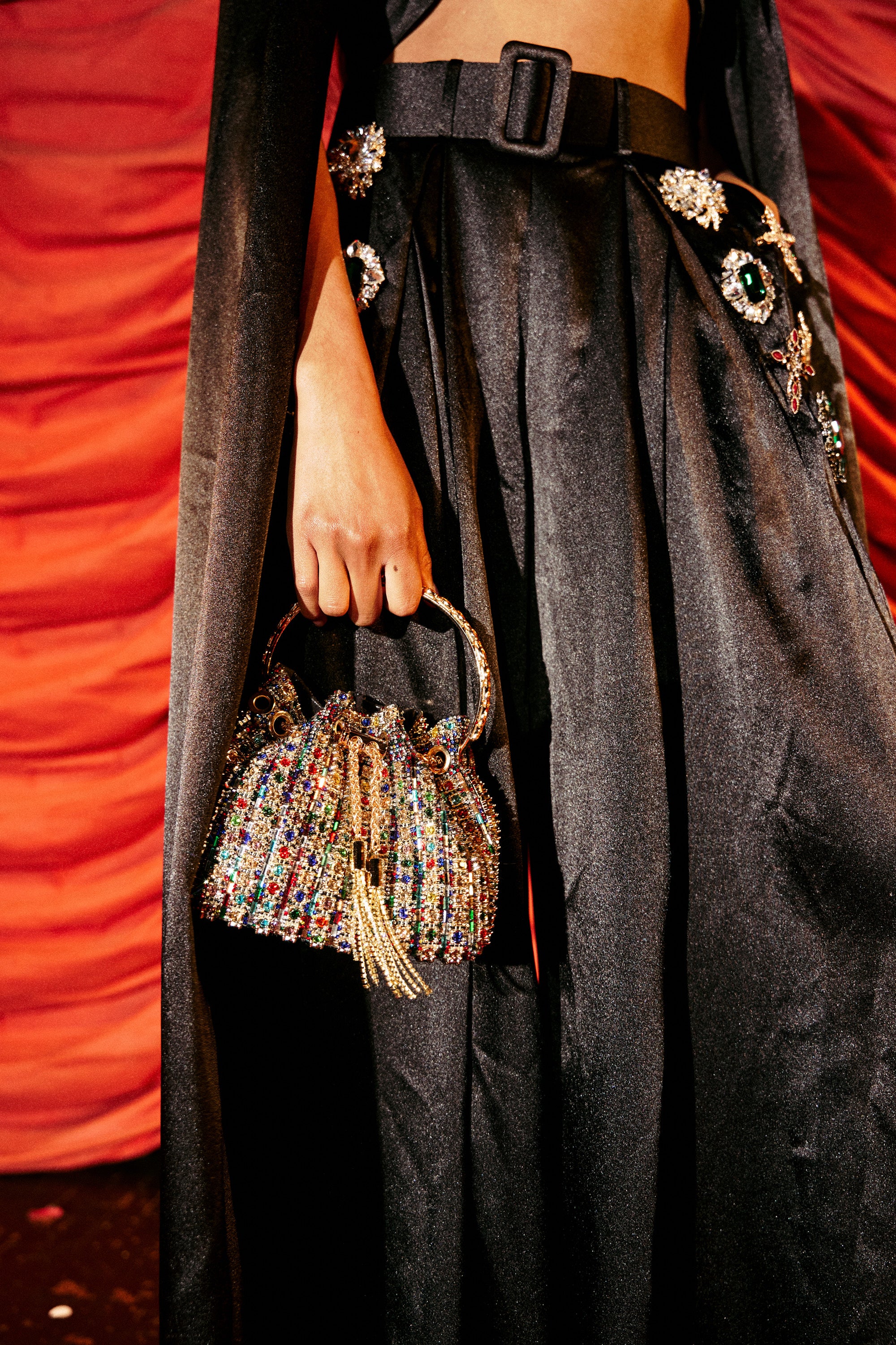 Holding the GEGE BLING PURSE, a person in chic black high-waisted jeweled pants stands before red draped fabric.