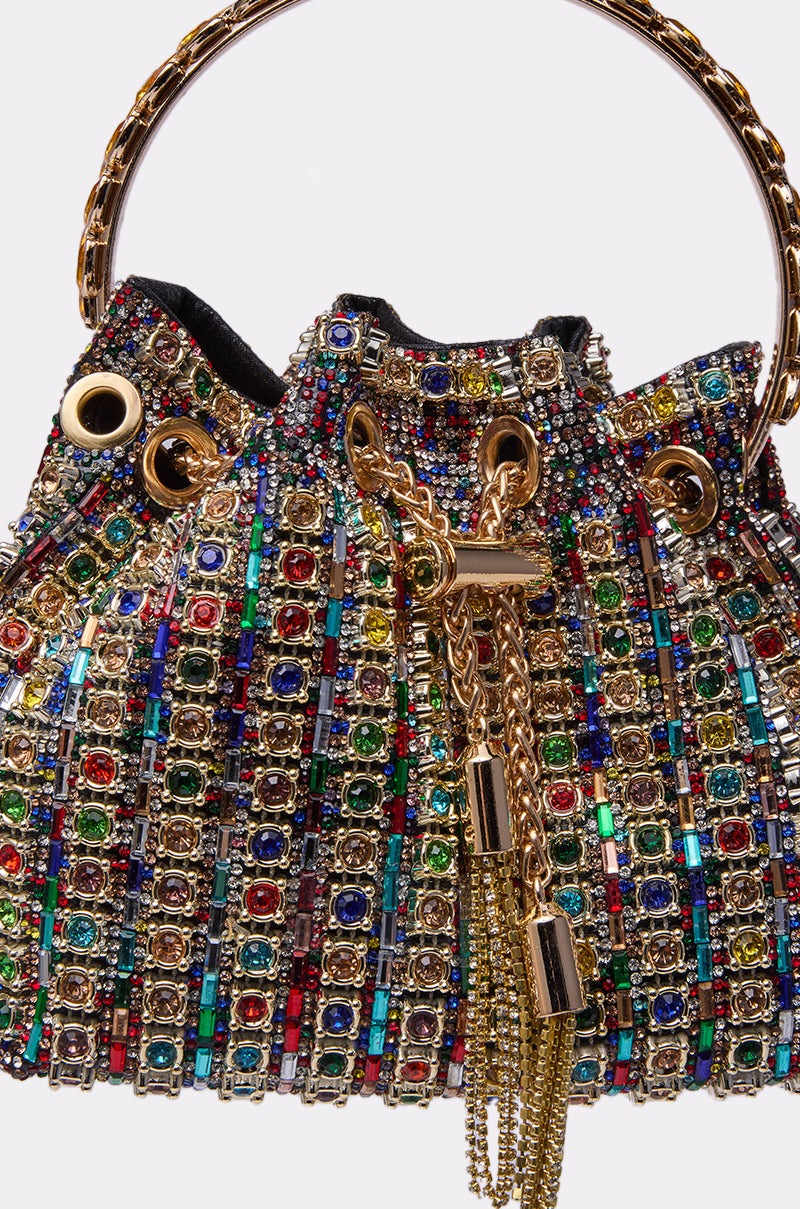 A close-up of the GEGE BLING PURSE, a luxury handbag adorned with multicolored gemstones in gold settings, featuring a gold chain drawstring, a gold circular handle, and an intricate rhinestone pattern.