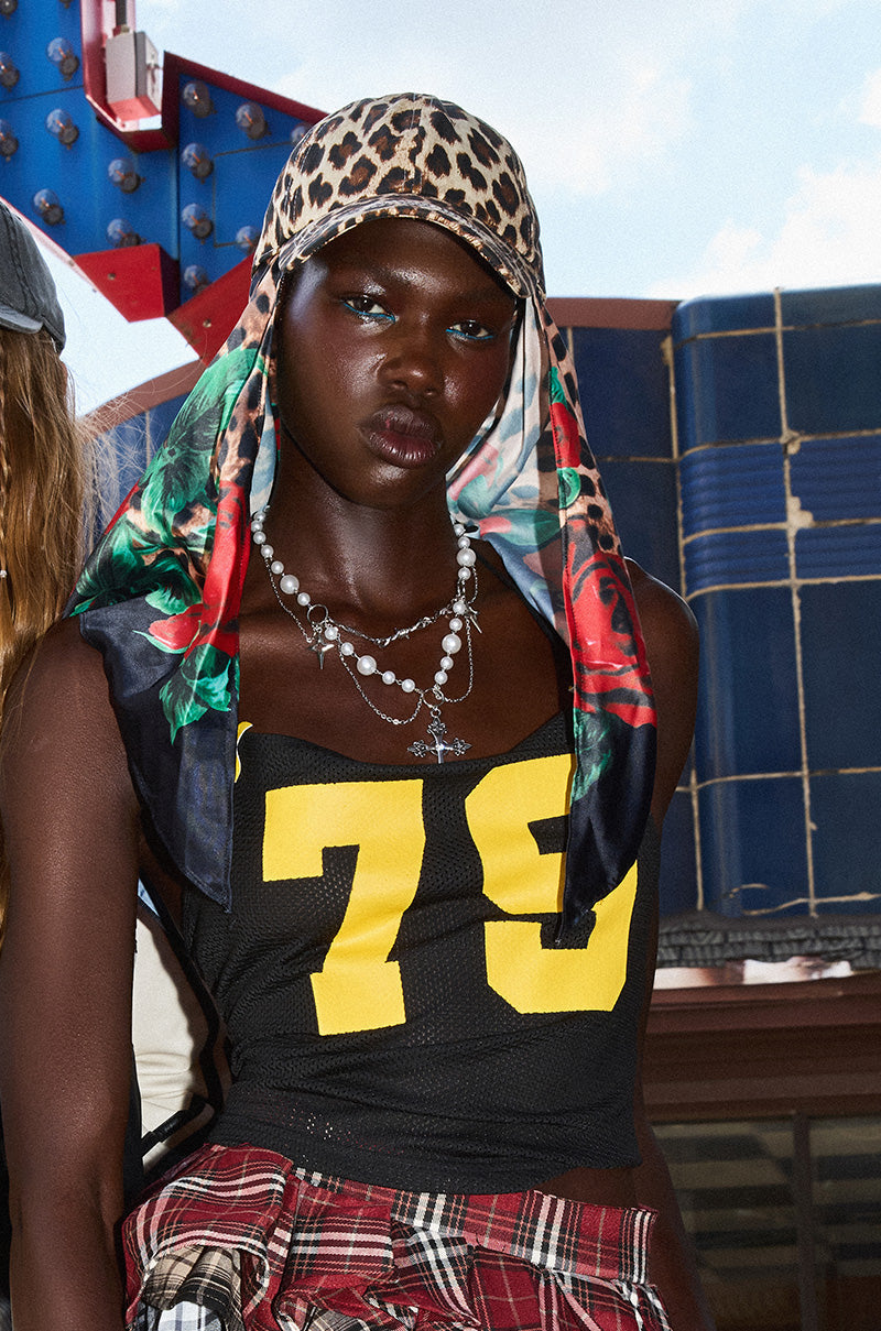 Wearing a black tank top with a yellow "79," layered necklaces, a plaid shirt at the waist, and the DOLCE LOVE HAT AND SCARF, someone stands outside a retro blue and red building. 0