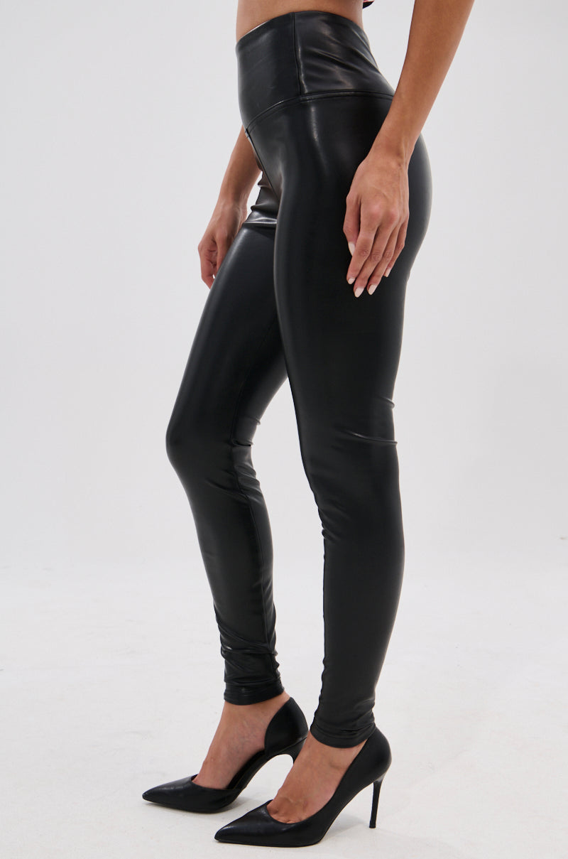 A person wears the SLIM THICC 4 WAY STRETCH LEGGING IN BLACK with black high-heeled shoes, standing on a white background. Only the lower body is visible, highlighting the sleek bodycon fit of these faux leather high waist leggings.