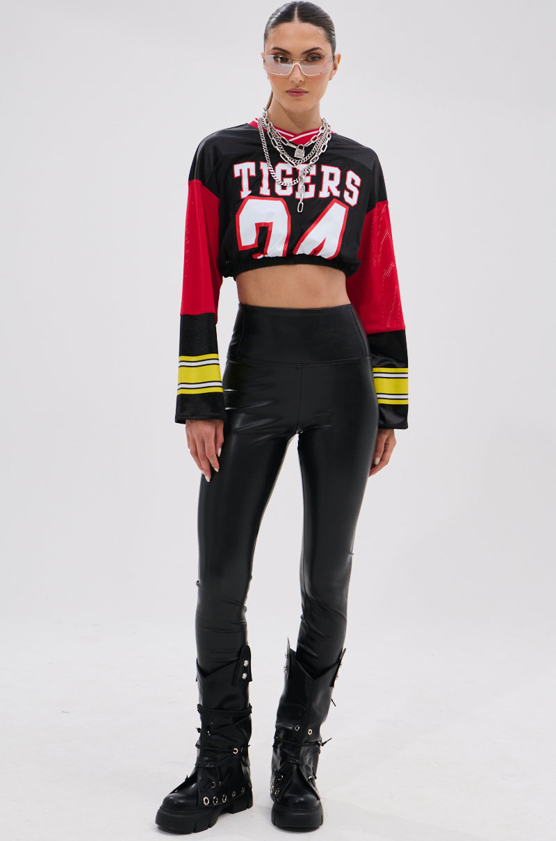A woman in a cropped "TIGERS 24" jersey, SLIM THICC 4 WAY STRETCH LEGGING IN BLACK, black combat boots, layered necklaces, and clear glasses stands confidently against a light background.