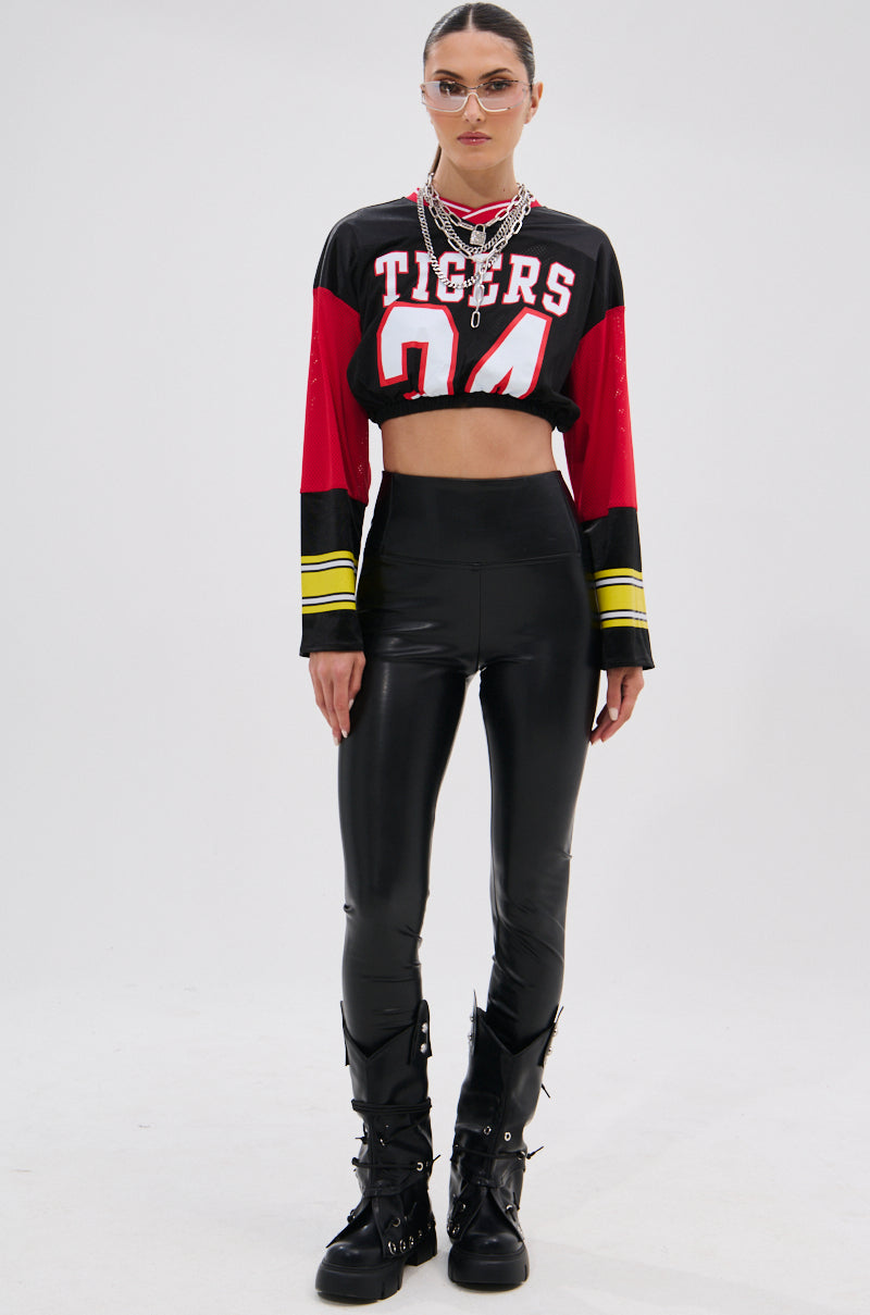 A woman wearing a cropped "TIGERS 94" jersey, SLIM THICC 4 WAY STRETCH LEGGING IN BLACK, chunky black boots with buckles, layered necklaces, and sunglasses stands against a plain white background.