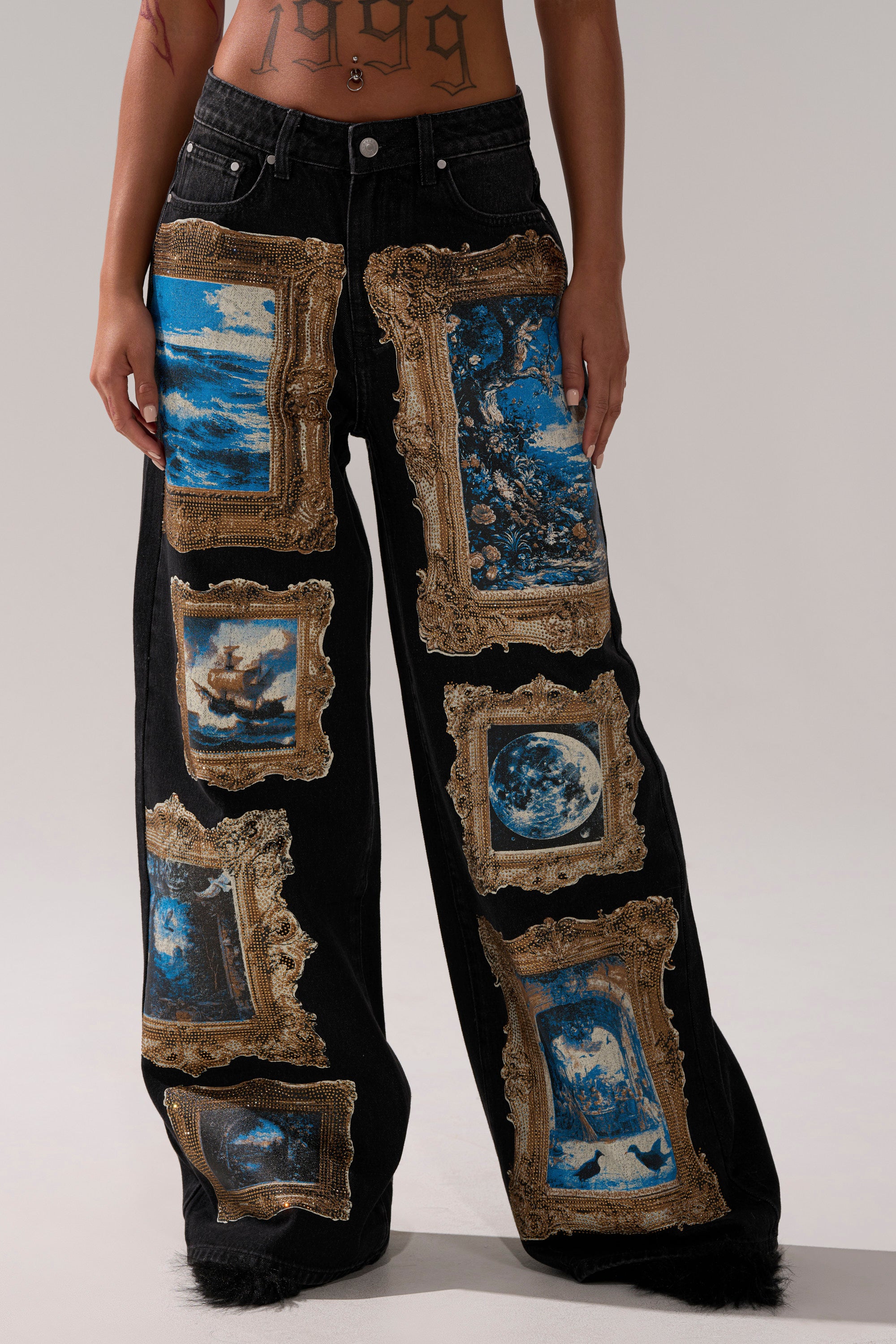 Wearing the WORK OF ART GRAPHIC WIDE LEG DENIM, featuring high-rise, gold-embroidered frames and blue artwork patches, a person’s lower torso and legs are shown against a plain background.