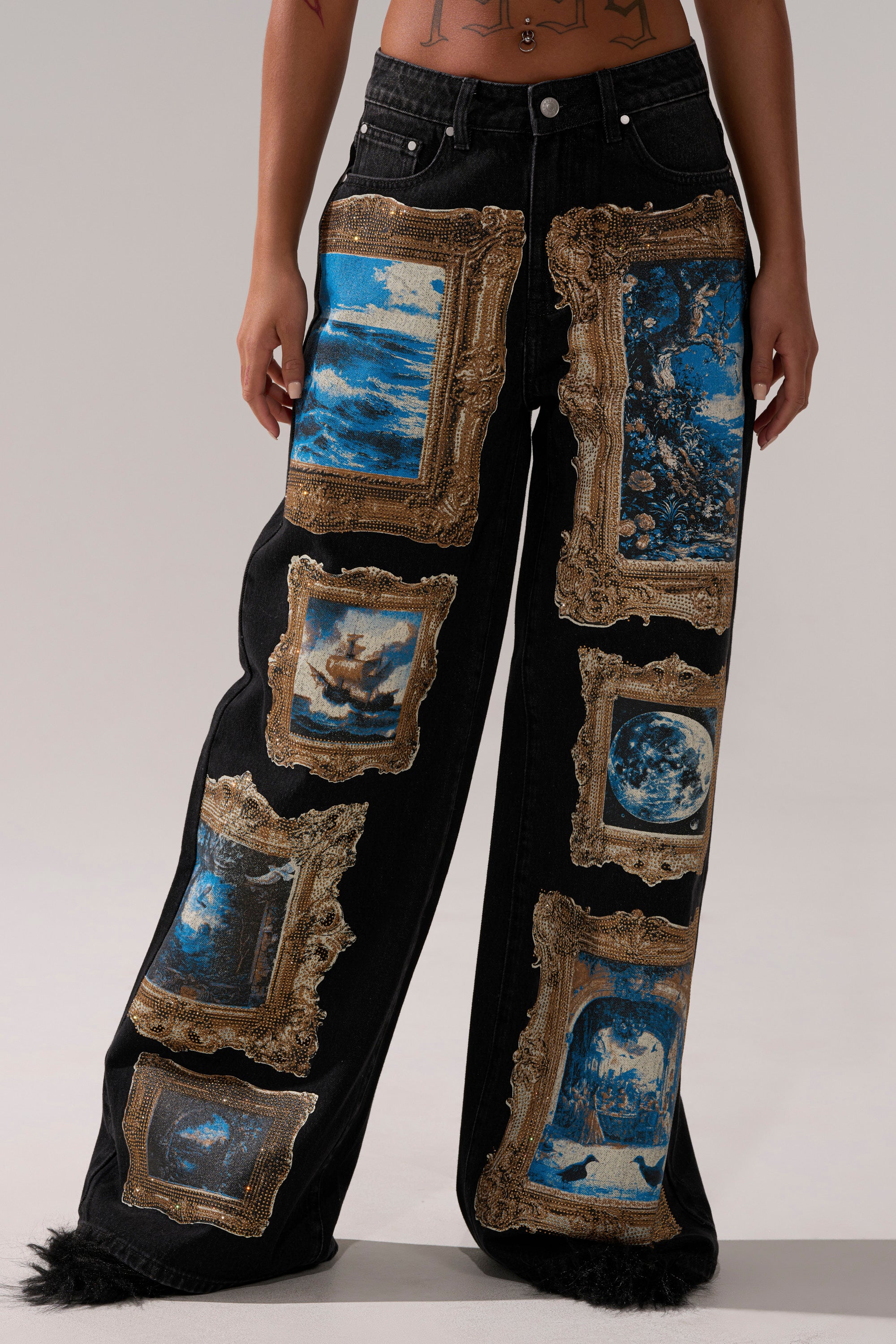 Person wearing WORK OF ART GRAPHIC WIDE LEG DENIM—high-rise, wide-leg jeans featuring gold embroidered frames with blue-toned art graphics like landscapes and celestial scenes—standing against a plain background.