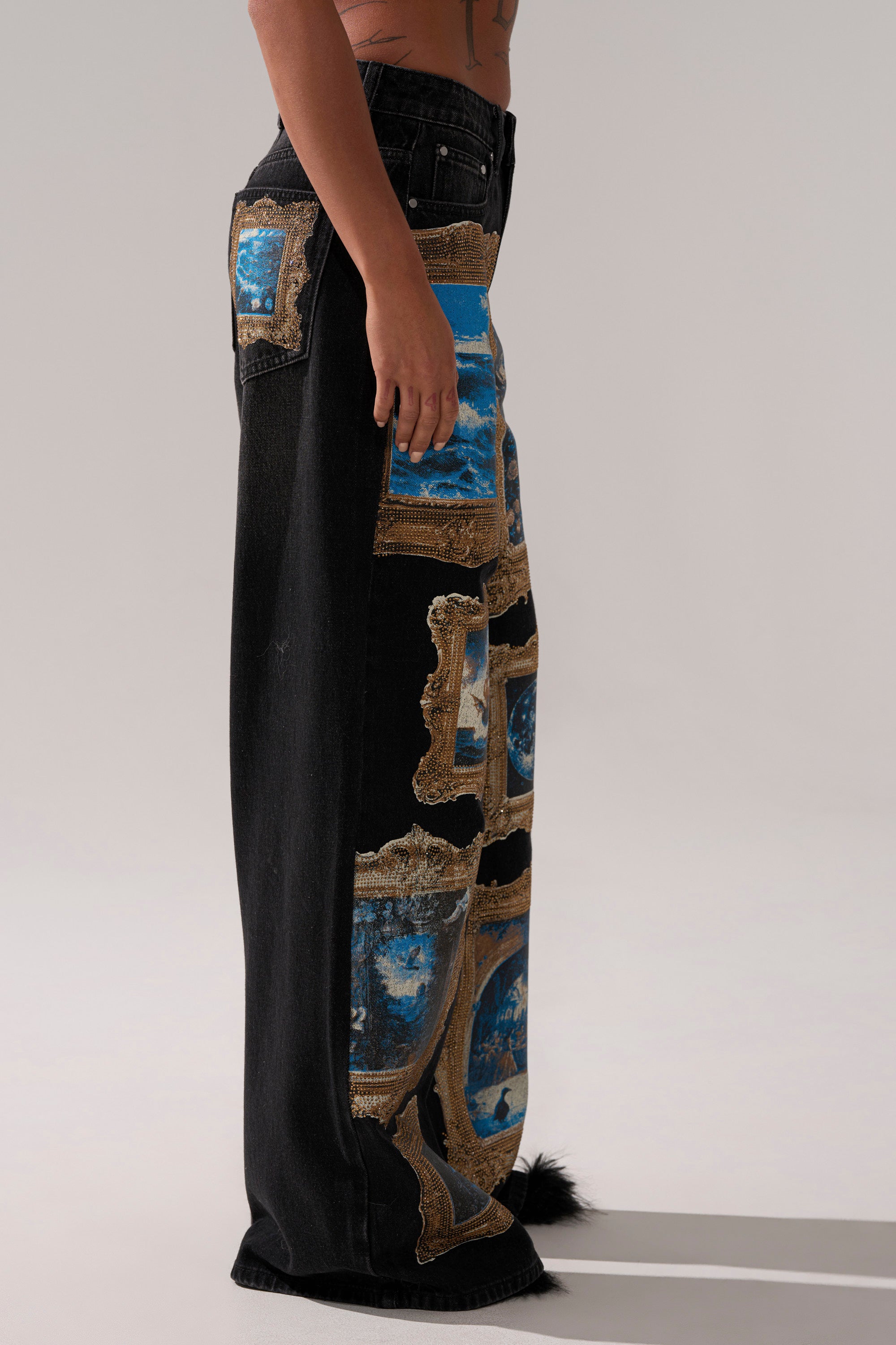 A person wears the WORK OF ART GRAPHIC WIDE LEG DENIM, featuring black wide-leg jeans with blue and gold tapestry-style patches resembling framed art. The image captures a side view against a plain background.