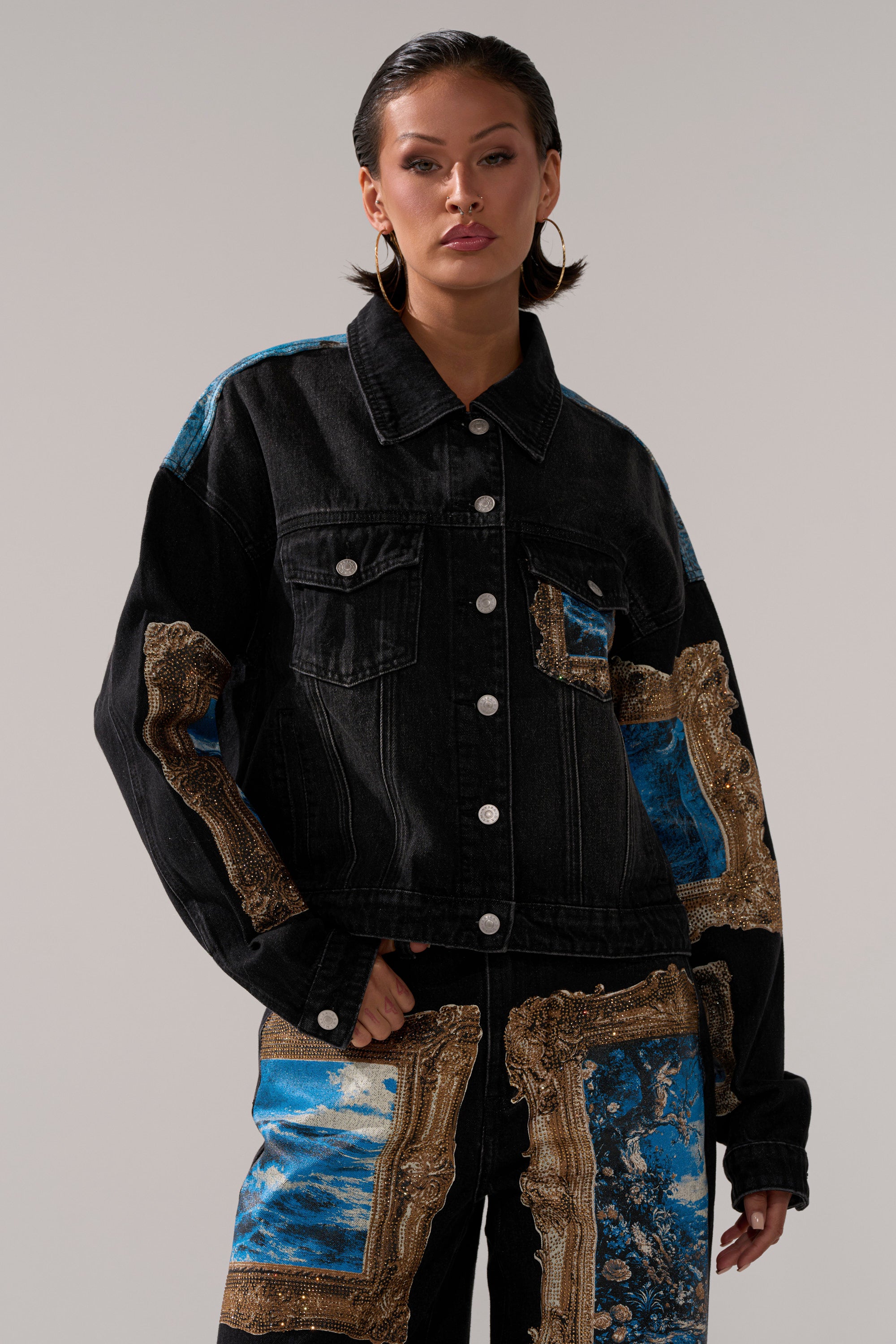 A person wears the WORK OF ART GRAPHIC DENIM JACKET, featuring gold and blue decorative patches, rhinestone trim, and matching pants, standing confidently against a plain background.