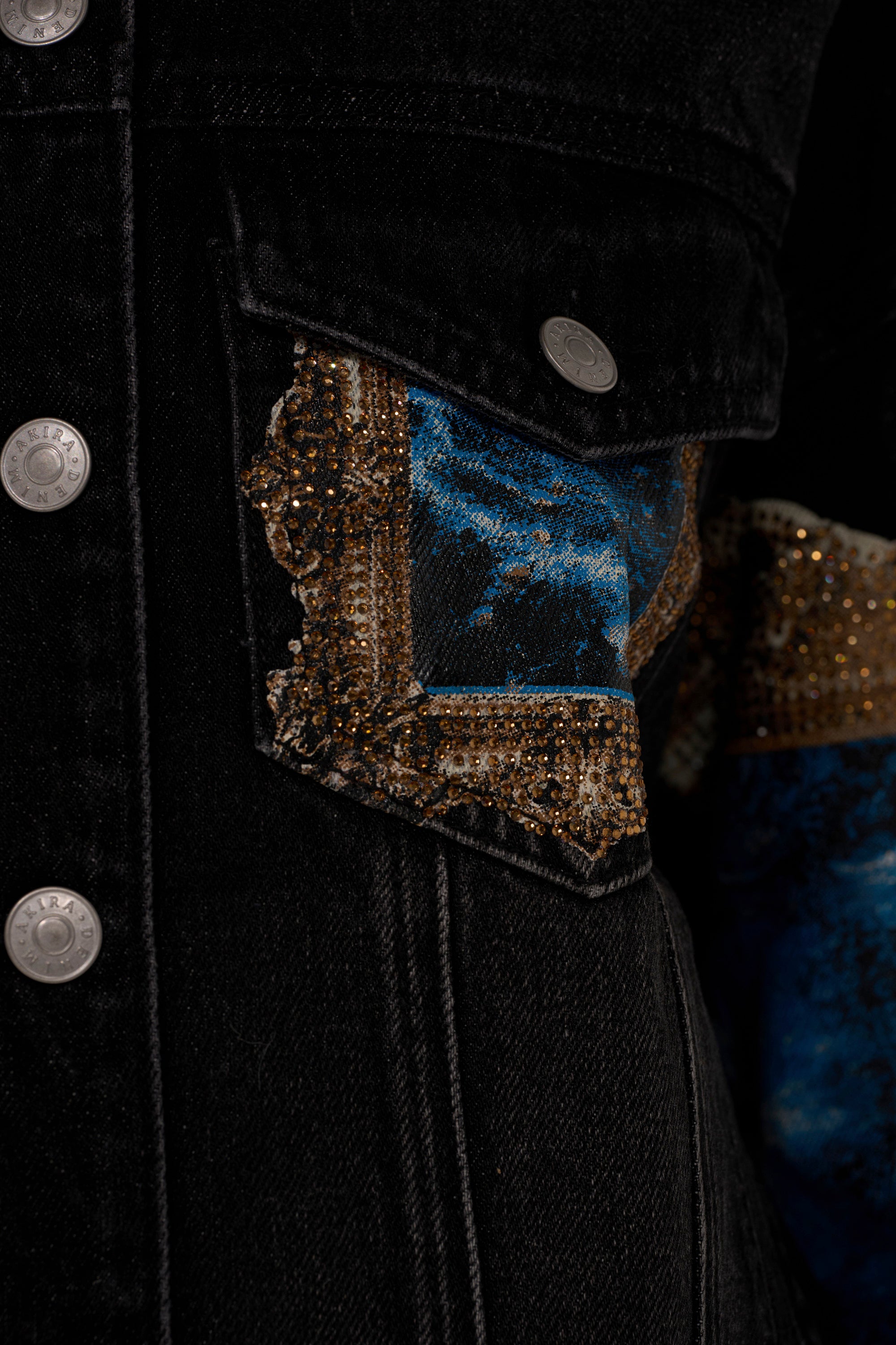 Close-up of the WORK OF ART GRAPHIC DENIM JACKET, a black denim jacket with silver buttons and a pocket featuring decorative blue and brown patterned fabric with gold beading detail peeking from beneath the flap.