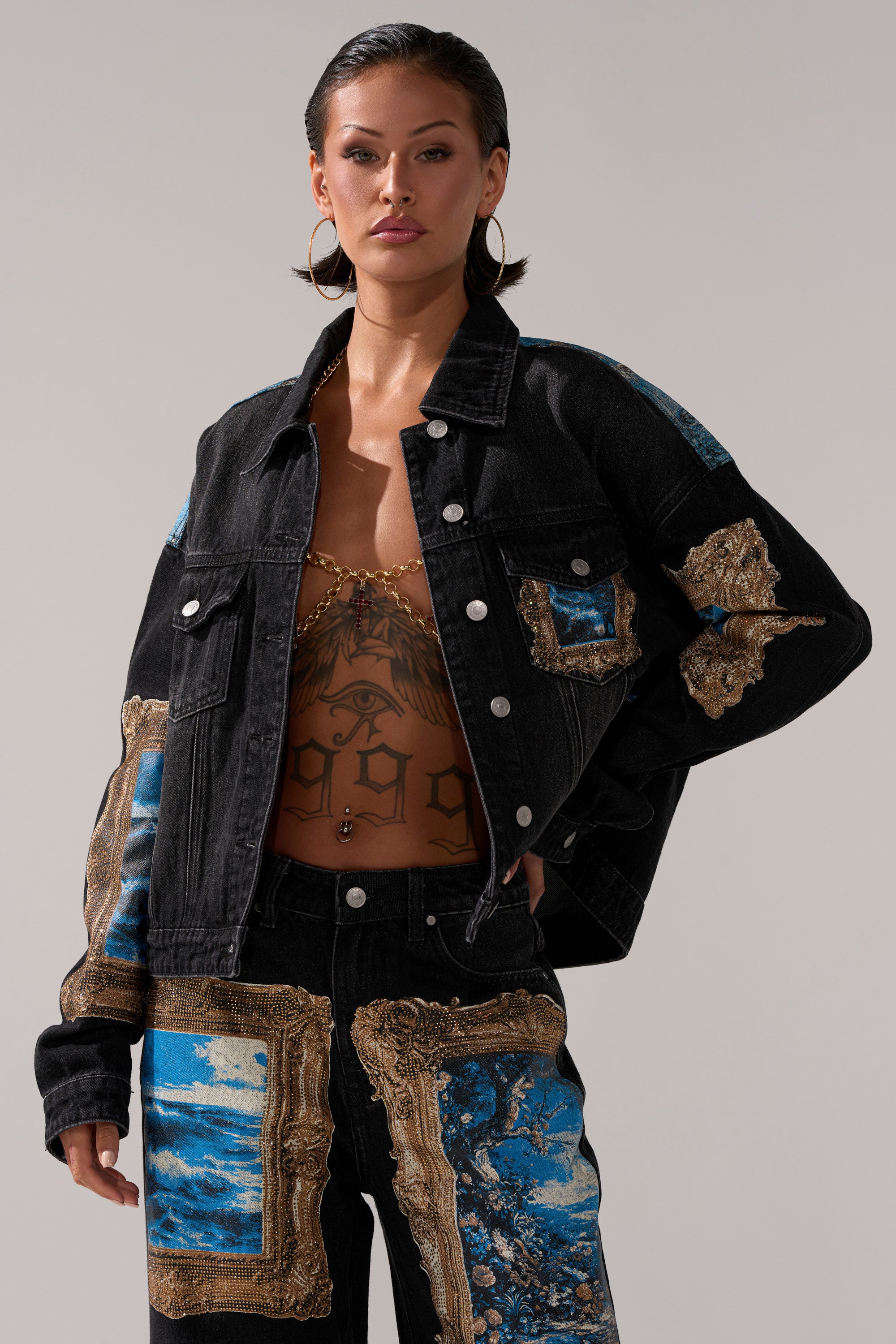 A confident woman showcases the WORK OF ART GRAPHIC DENIM JACKET and matching pants with blue and gold patchwork. She adds bold flair with large hoop earrings and a chest tattoo, standing against a plain, light-colored background.
