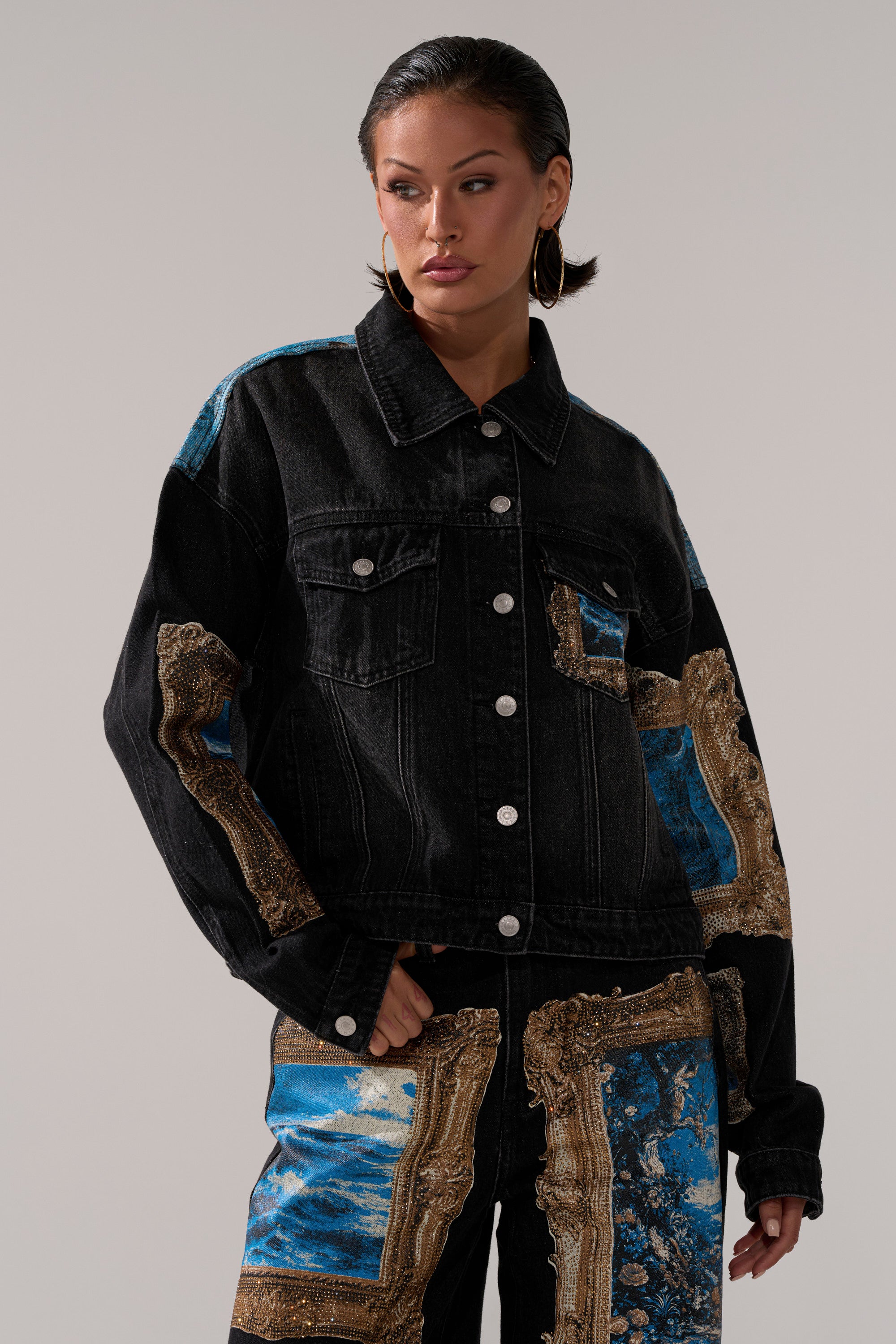 A person in the WORK OF ART GRAPHIC DENIM JACKET, featuring ornate gold and blue baroque-style patterns with matching pants, stands against a neutral background, looking to the side.