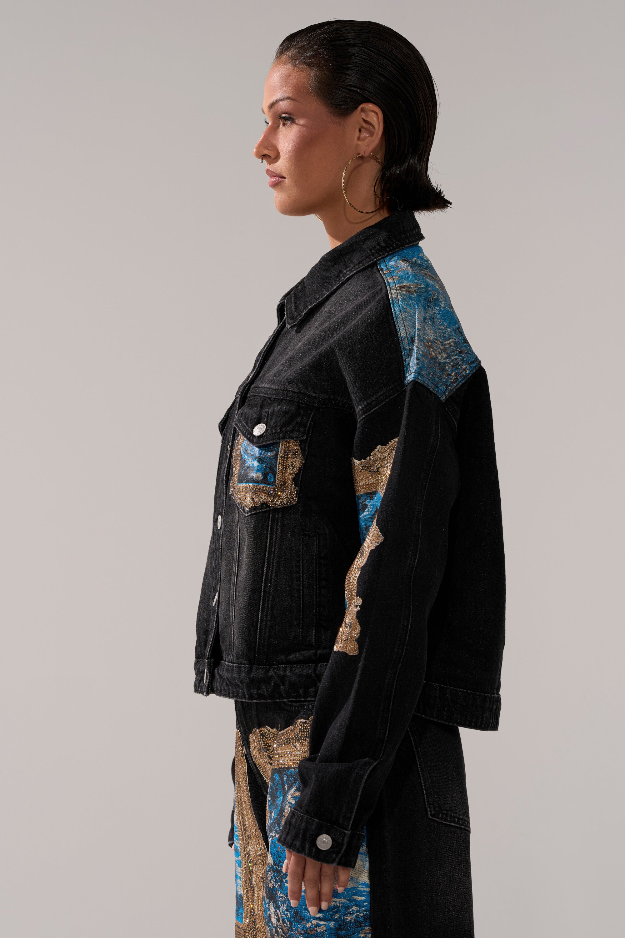 A person with slicked-back hair and large hoop earrings stands in profile against a plain background, wearing the WORK OF ART GRAPHIC DENIM JACKET with blue and gold patterned patches.
