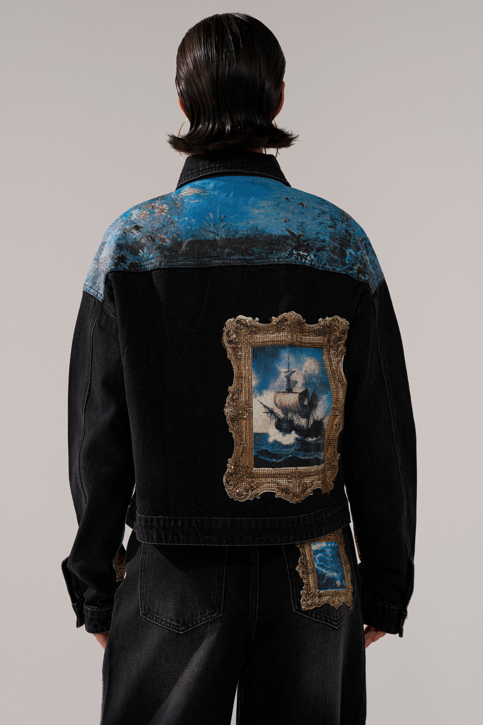 A person with dark, slicked-back hair wears the WORK OF ART GRAPHIC DENIM JACKET—black denim with blue details and a patch resembling a framed ship painting—and matching jeans featuring a similar patch on the back pocket.