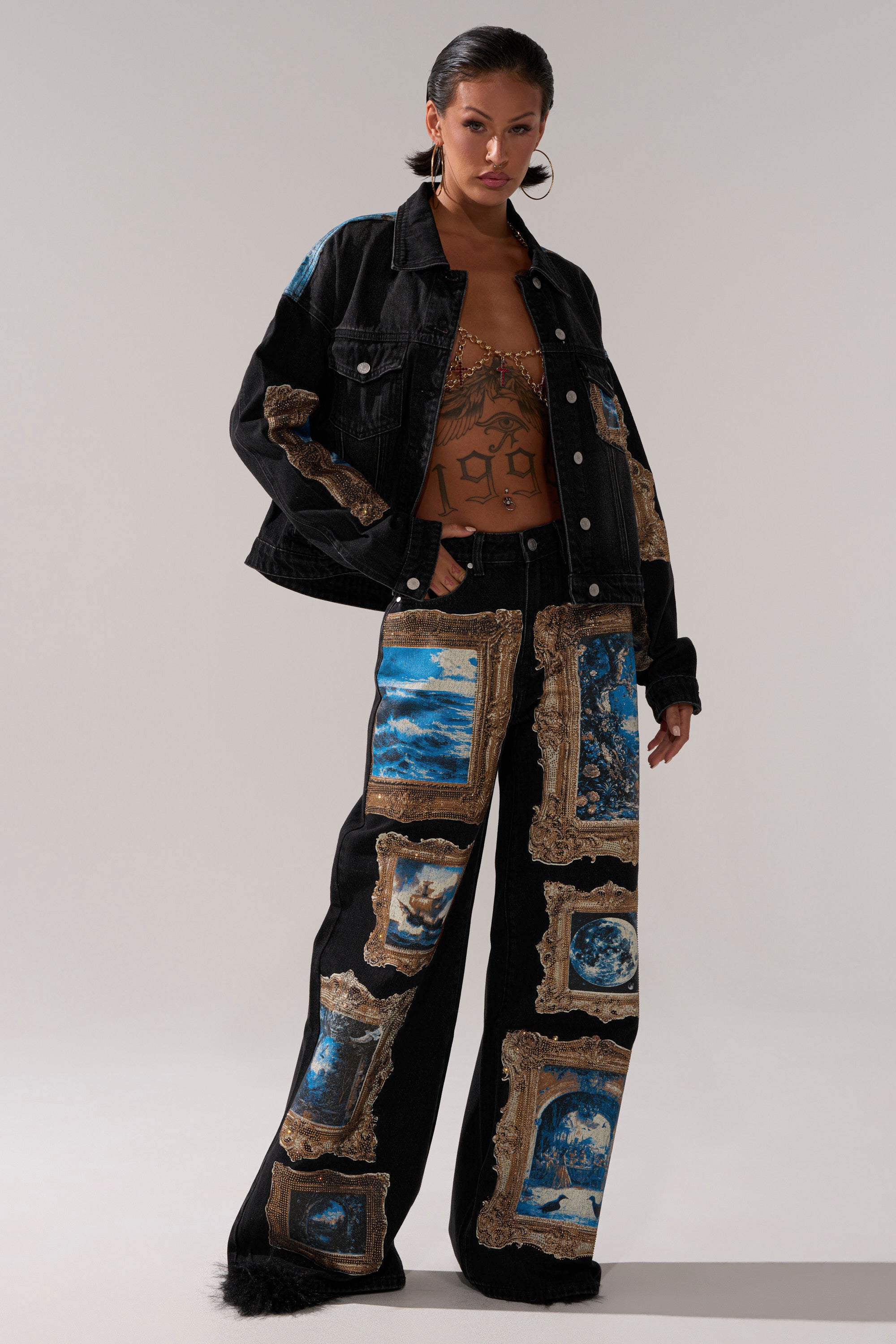 A woman wears the WORK OF ART GRAPHIC DENIM JACKET, featuring rhinestone trim and blue and gold framed artwork prints, styled with matching wide-leg pants, slicked-back hair, and a brown graphic crop top underneath.