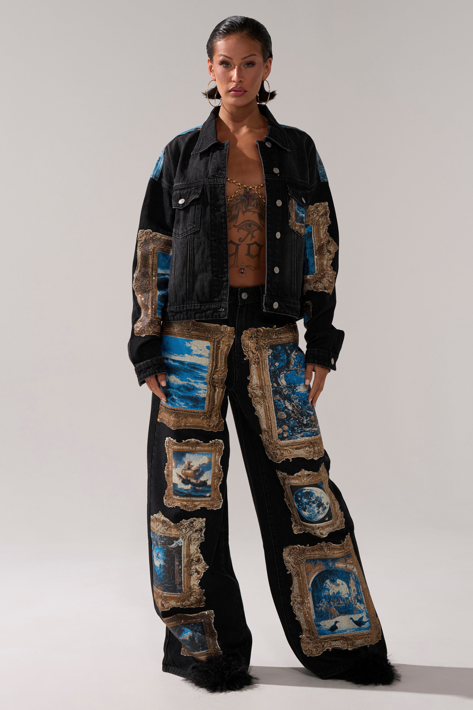 A woman with slicked-back hair and visible chest tattoos faces forward, wearing the WORK OF ART GRAPHIC DENIM JACKET adorned with blue and white picture frame graphics, paired with matching wide-leg pants.