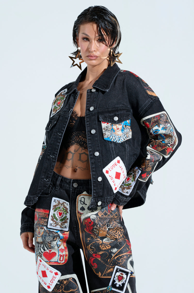 Against a plain white background, a person confidently wears a QUEEN OF HEARTS PRINTED AND EMBELLISHED DENIM JACKET with matching pants featuring vibrant playing card designs. Large star-shaped earrings enhance their look while a tattoo adorns their midriff.
