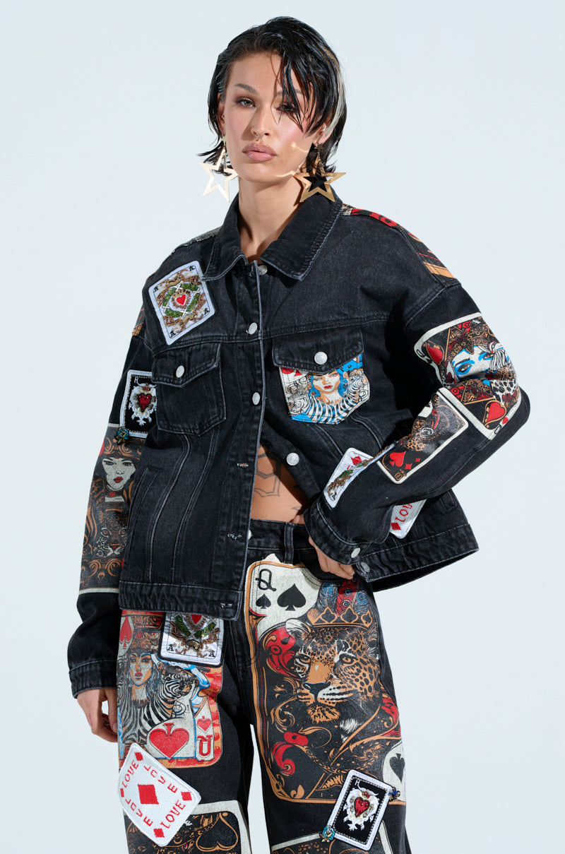 A person exudes charisma in the Queen Of Hearts Printed and Embellished Denim Jacket, with colorful playing card graphics. With short hair and large star-shaped earrings, they confidently channel the jacket's bold spirit.