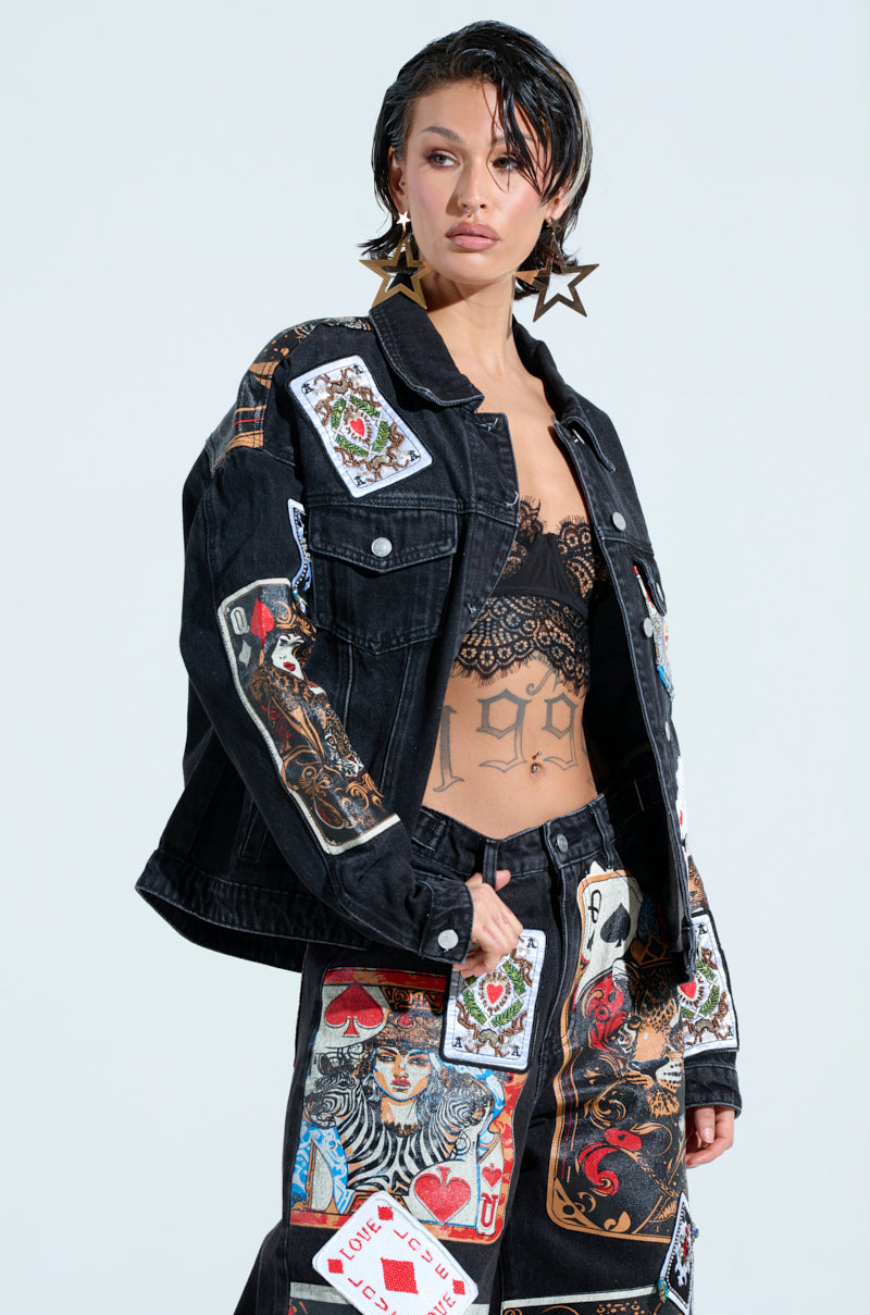A person poses confidently in a bold QUEEN OF HEARTS PRINTED AND EMBELLISHED DENIM JACKET and pants, featuring playful card graphics. They sport short, wet-look hair, large star-shaped earrings, and a visible stomach tattoo against a plain light background.