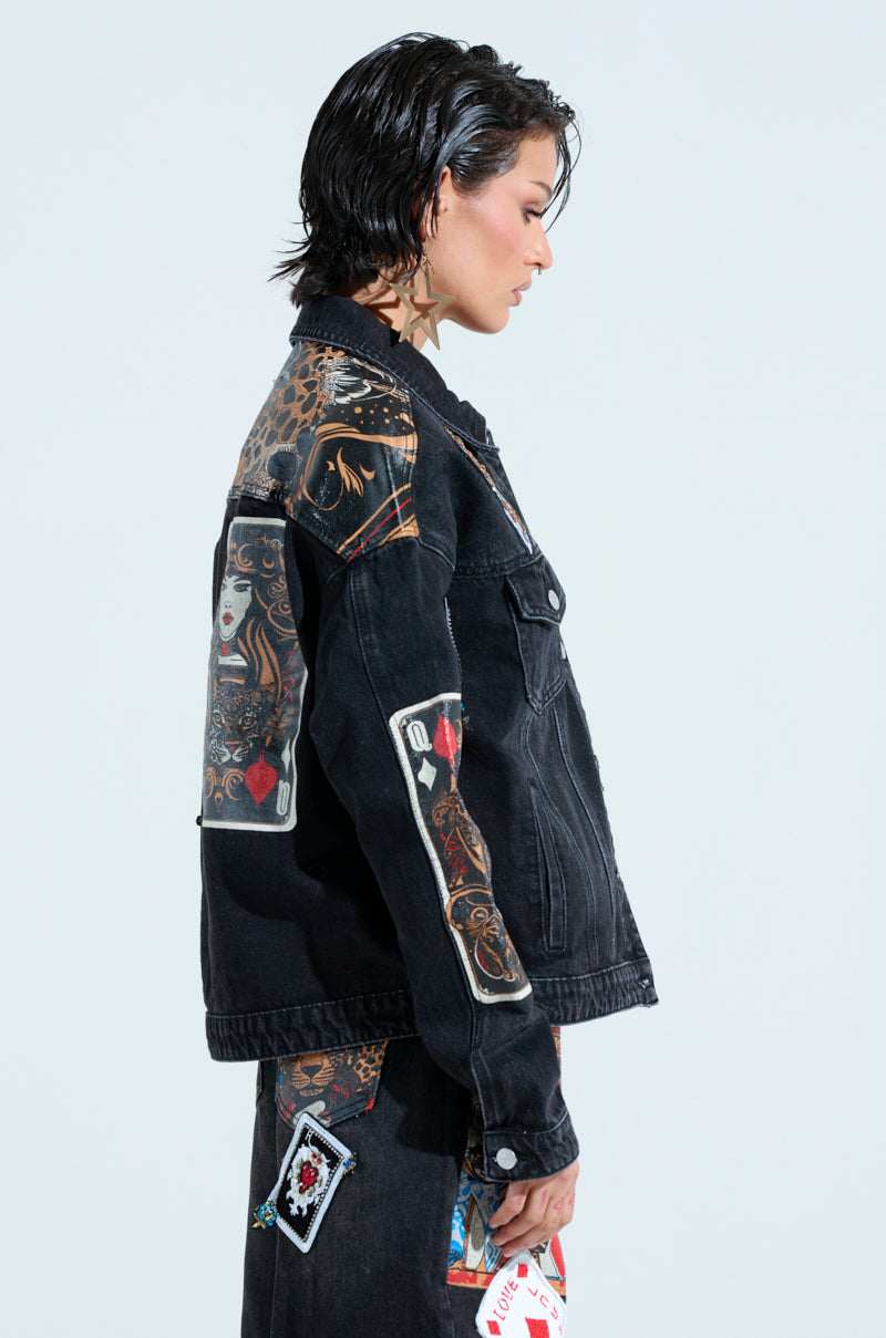 Wearing the QUEEN OF HEARTS PRINTED AND EMBELLISHED DENIM JACKET and a matching skirt adorned with card designs, the individual showcases embroidered patches, a leopard print section, short wet-look hair, and star-shaped earrings for an edgy charm.