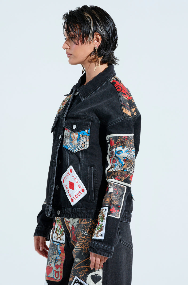 Wearing the QUEEN OF HEARTS printed and embellished denim jacket paired with pants adorned in vivid, artistic playing card graphics, including faces and designs. They face left, with hair styled back and wearing star-shaped earrings.