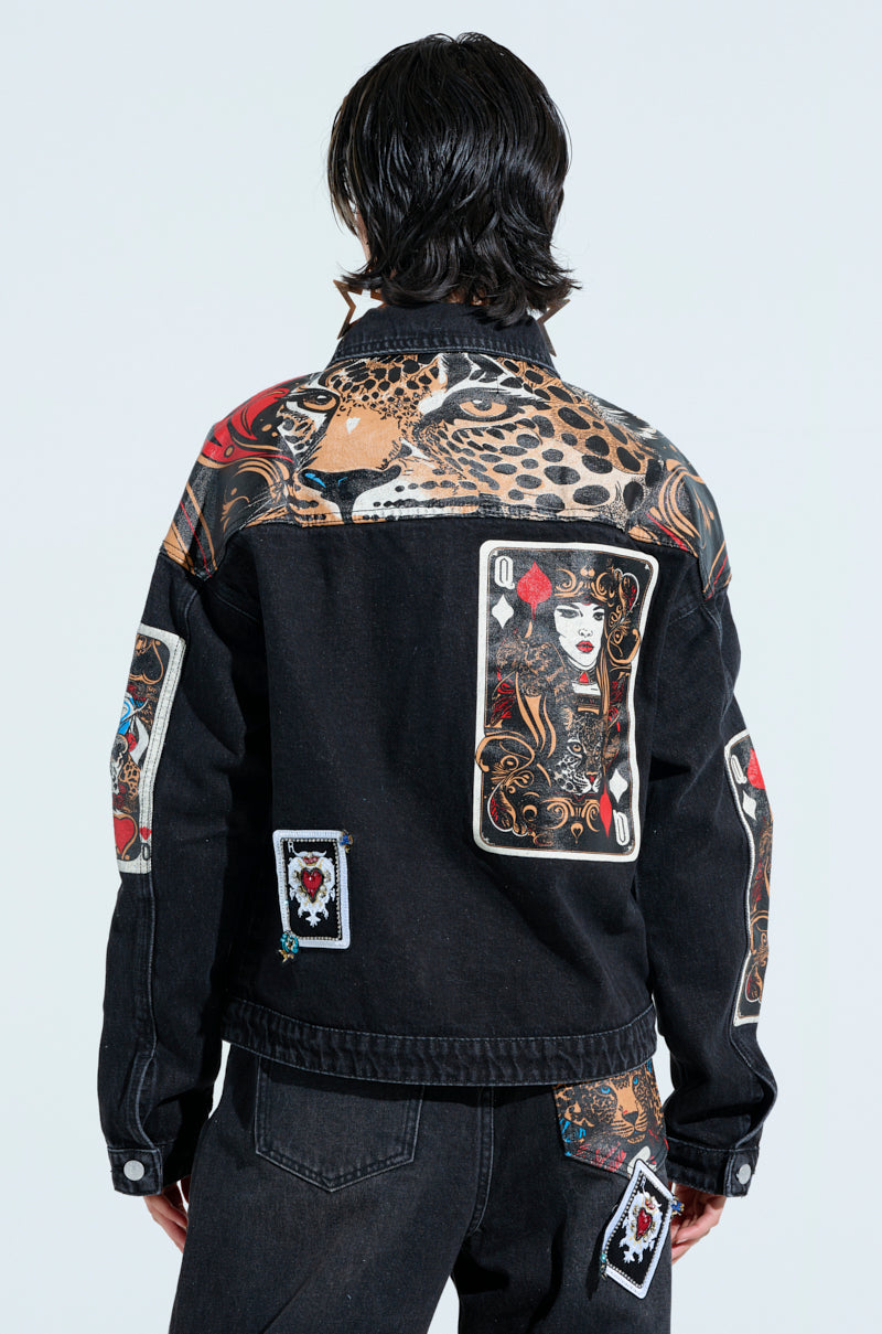 A person wears the Queen of Hearts printed and embellished denim jacket, featuring a large leopard print and card graphics with the Queen of Hearts on the back, paired with matching pants showcasing similar patterns for a bold look.
