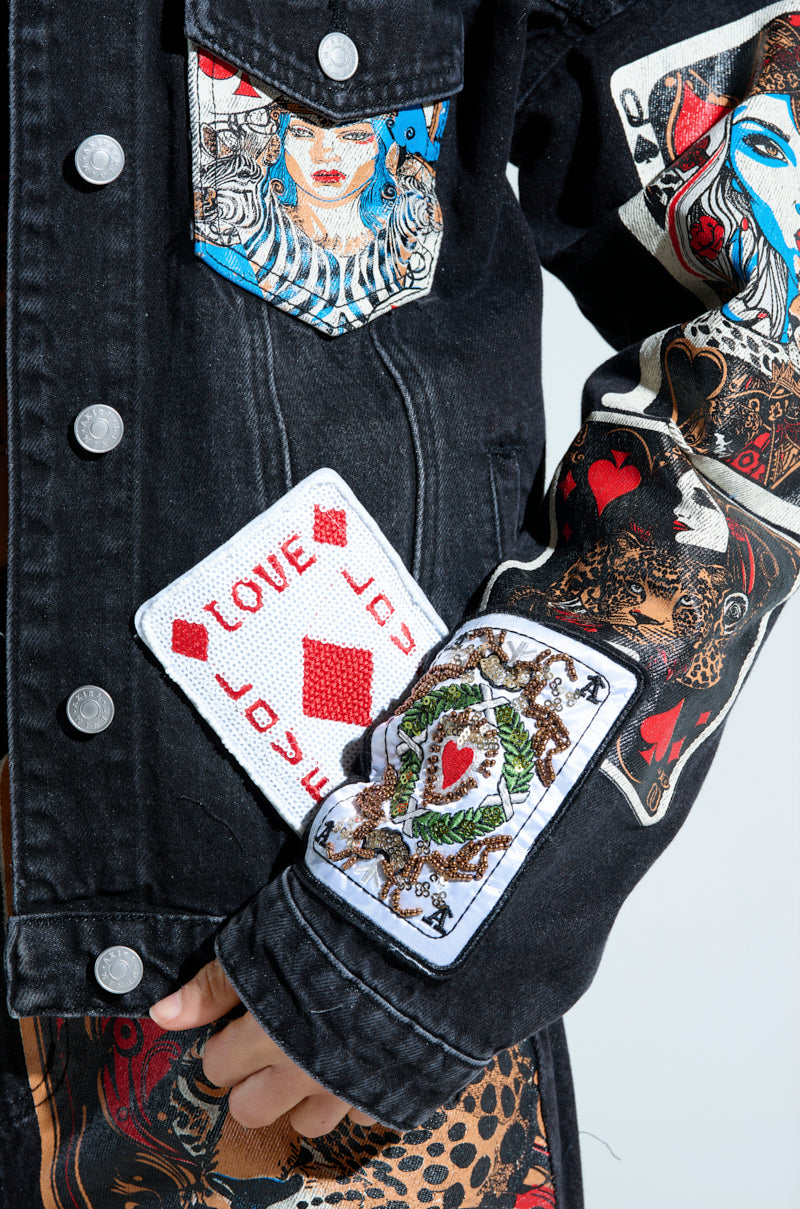 A person in a QUEEN OF HEARTS PRINTED AND EMBELLISHED DENIM JACKET, adorned with intricate card designs and colorful graphics, featuring queen, heart patches, and "LOVE" on the sleeve for a truly unique look.