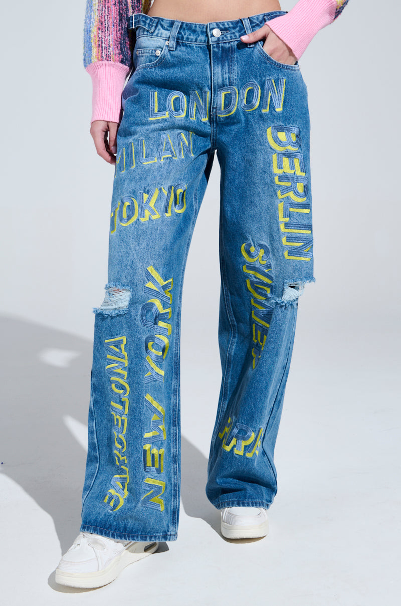 A person in Jetsetter Embroidery Wide Leg Jeans with yellow city names like London, Milan, Tokyo, Berlin, Barcelona, and New York. The knee-ripped jeans pair well with white sneakers and a pink multicolored sweater.