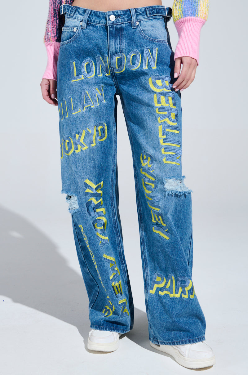 Someone is wearing JETSETTER EMBROIDERY WIDE LEG JEANS featuring bold yellow city names like "LONDON," "MILAN," and "TOKYO." They have a relaxed fit with knee rips. Paired with a colorful long-sleeved top and white shoes, these jeans offer a chic, urban vibe.